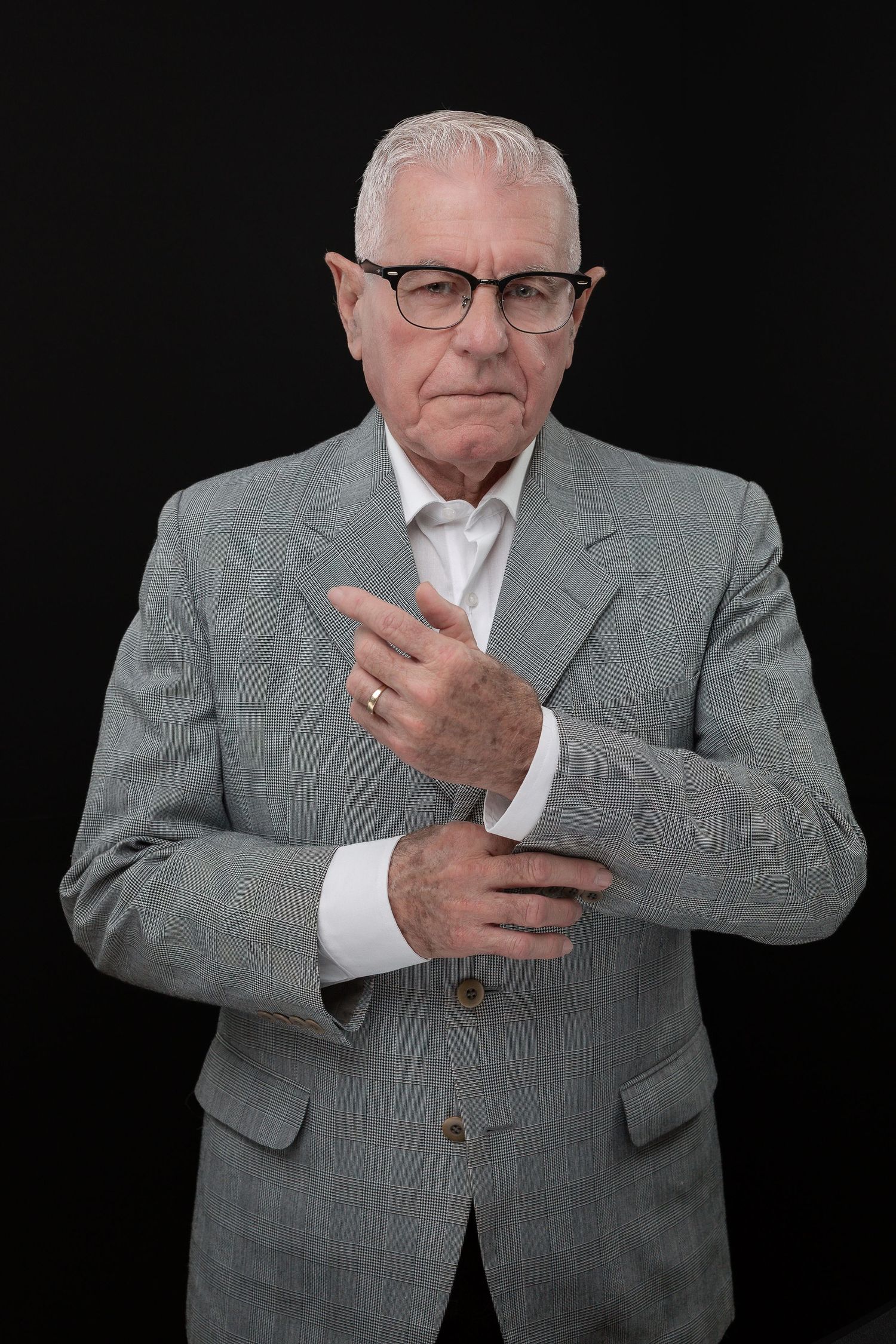 Senior executive in grey suit and glasses gesturing thoughtfully against dark background.