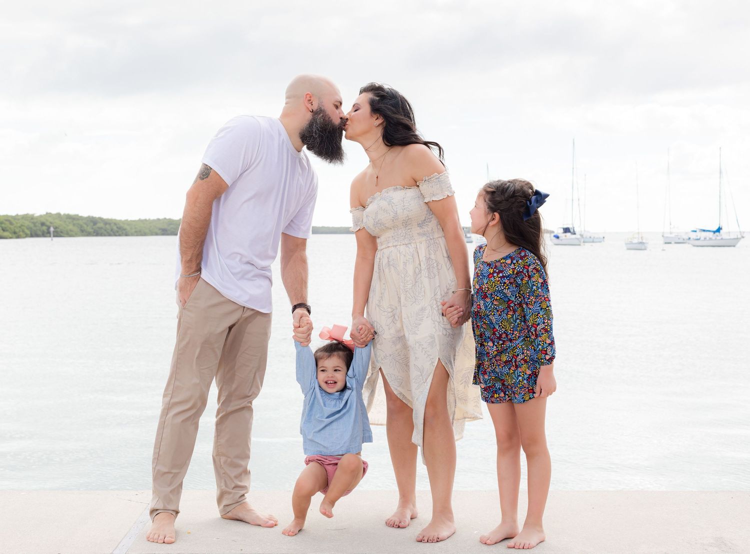 A family enjoys a playful moment together on a beach with boats visible in the background on a bright sunny day.