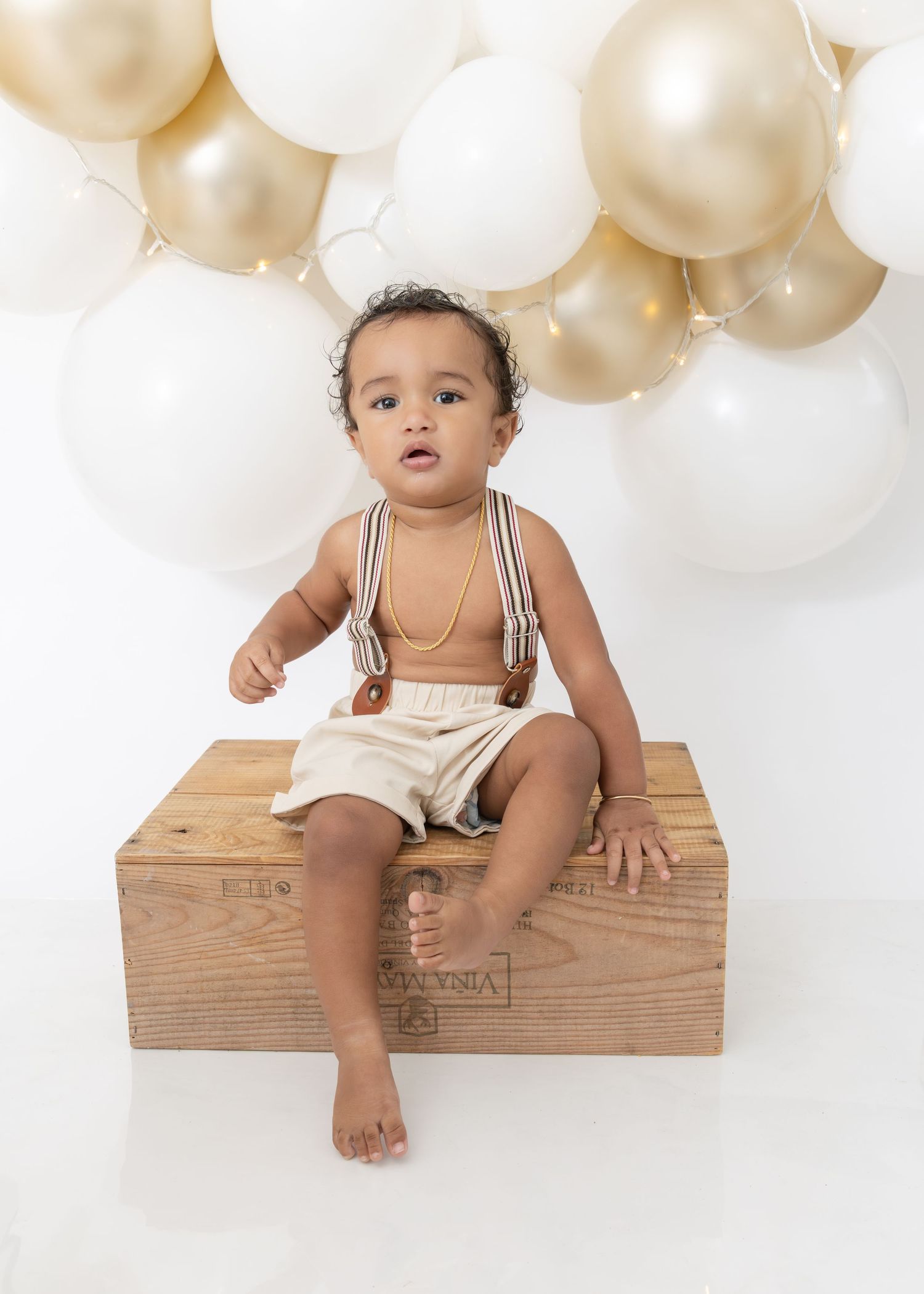 Baby in striped overalls sits on wooden crate against backdrop of white and gold balloons for milestone photo session.