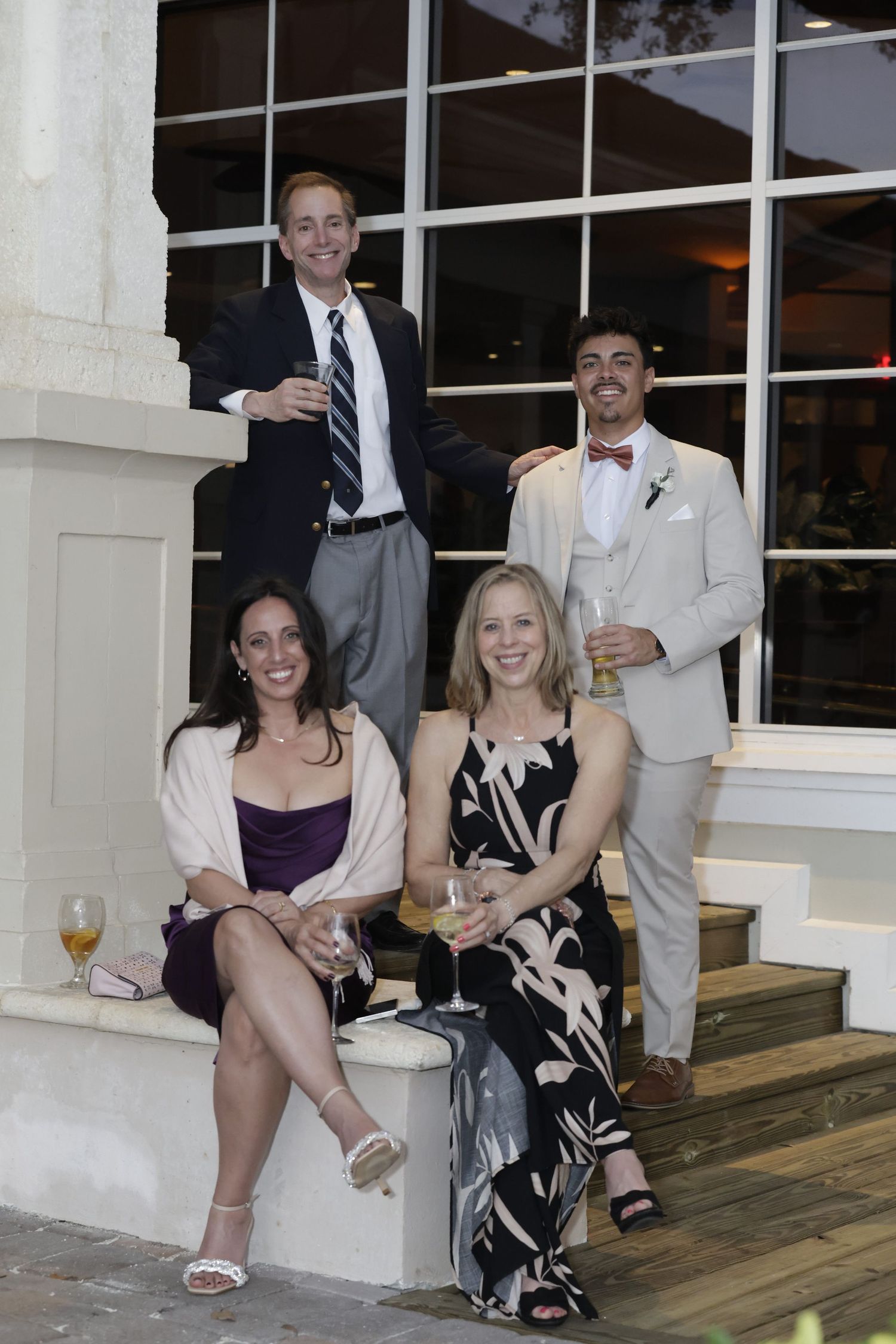 A group in formal attire sits casually on outdoor wooden decking during an evening event.