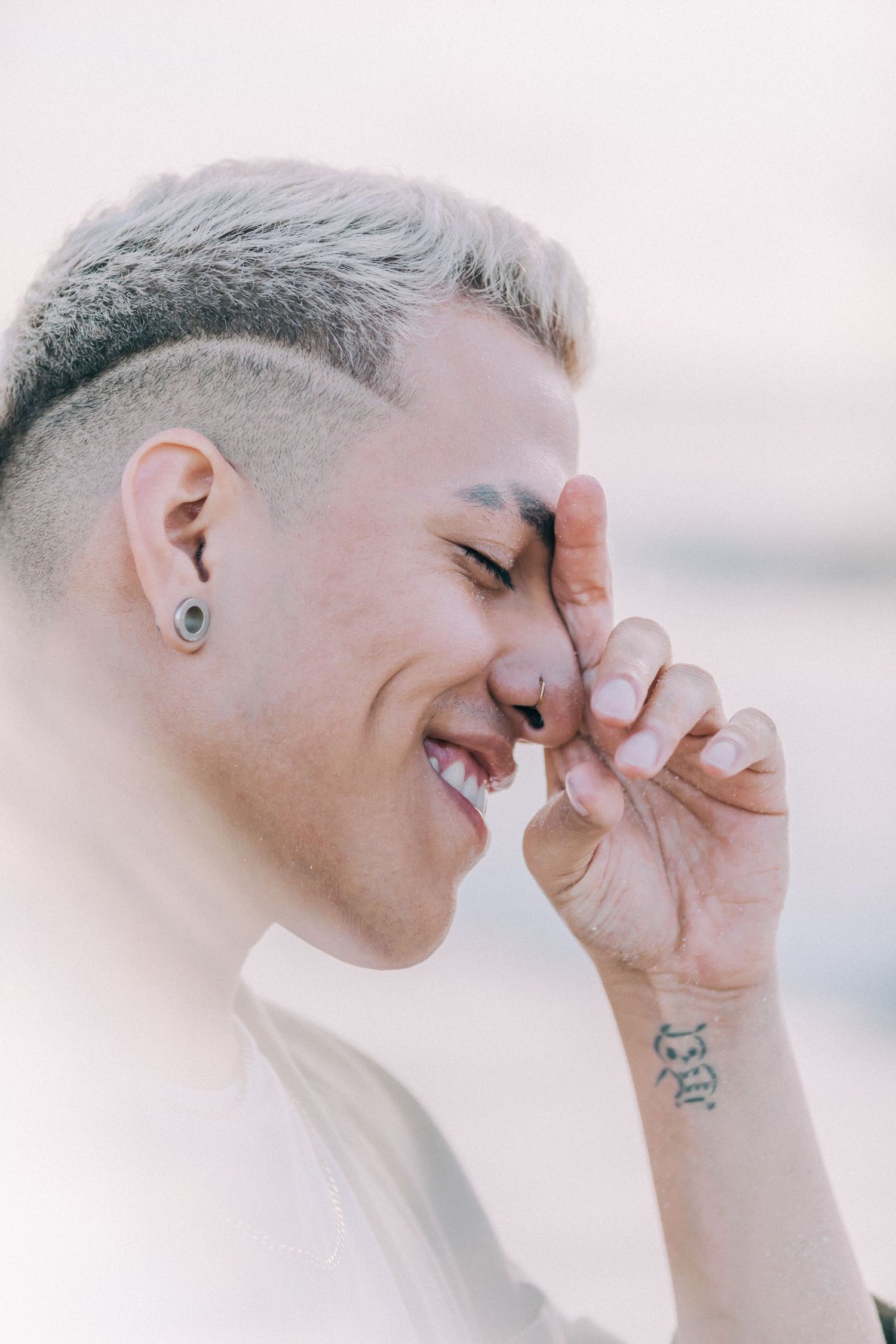 Close-up portrait of person with platinum hair and piercings smiling with hand near face.