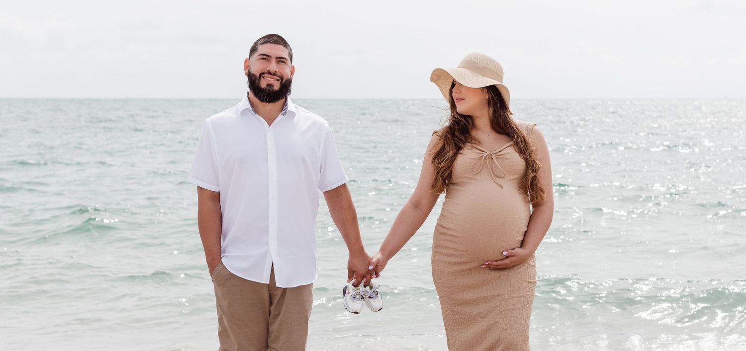 A couple holds hands while standing on a sunny beach, the woman wearing a beige maternity dress and sunhat.