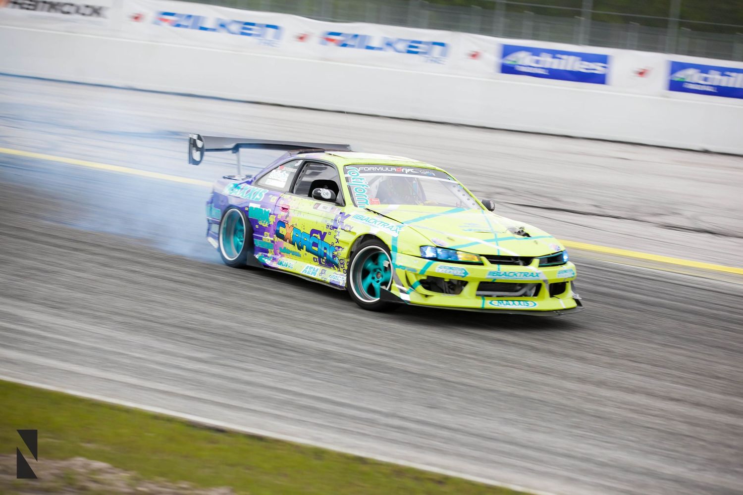 A green and turquoise drift car slides sideways on a racetrack creating smoke from its tires during competition.