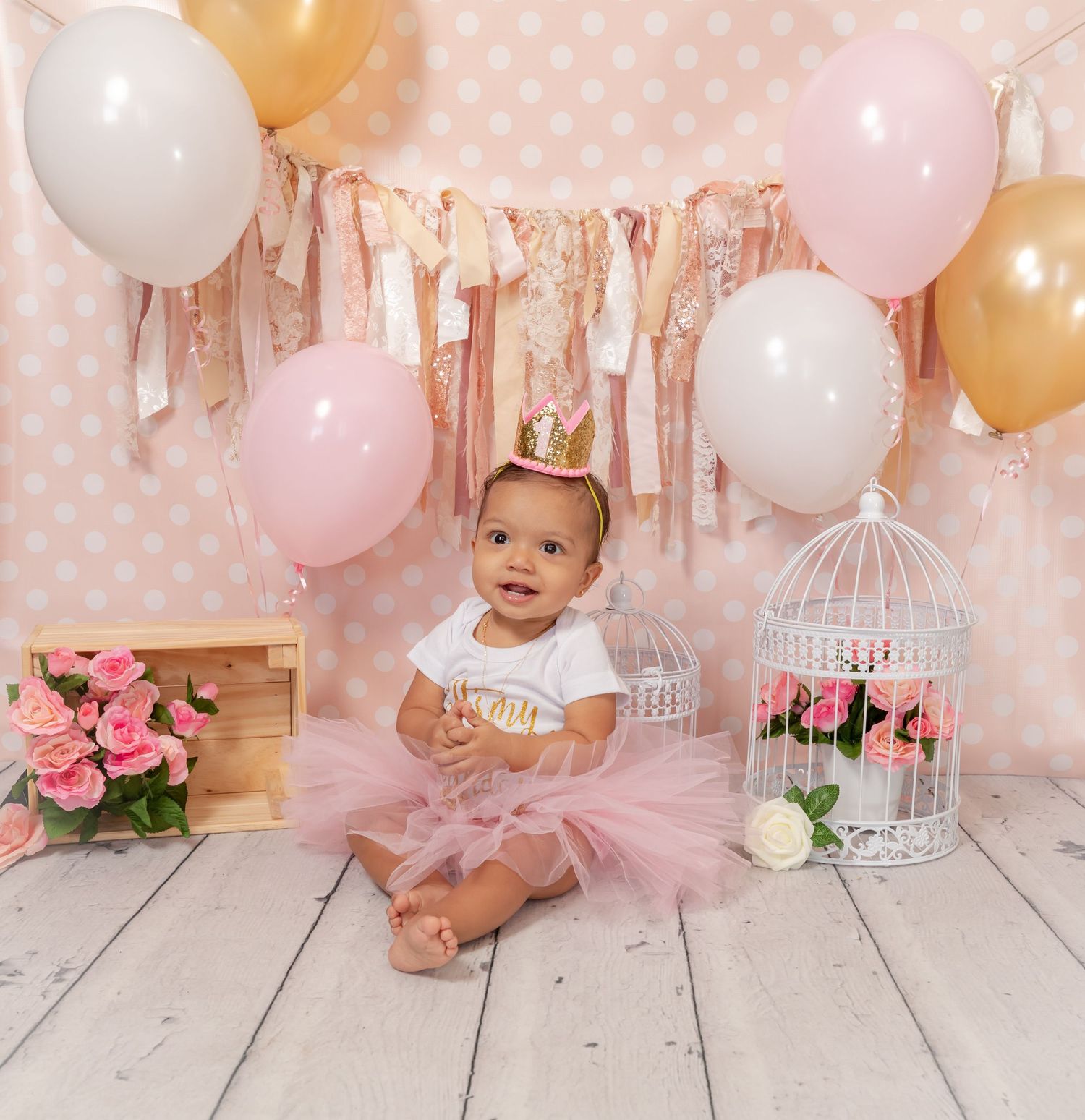 Party setup with pink and gold balloons, decorative banner, floral decorations, and white birdcage against polka dot backdrop.