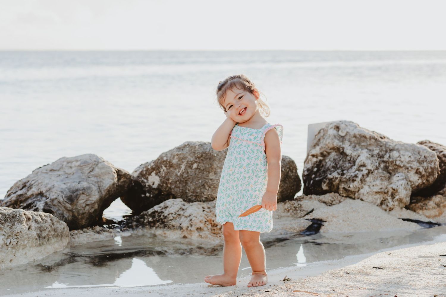 A child in a light blue dress poses near rocky formations at the beach.