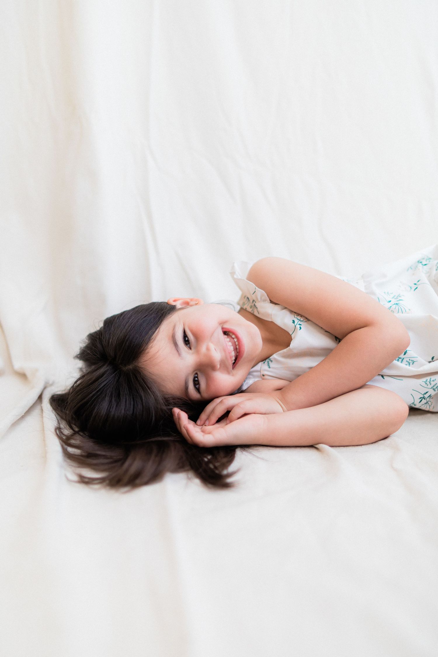 Young person lying down on white bedding wearing a floral dress smiling with dark hair spread out.