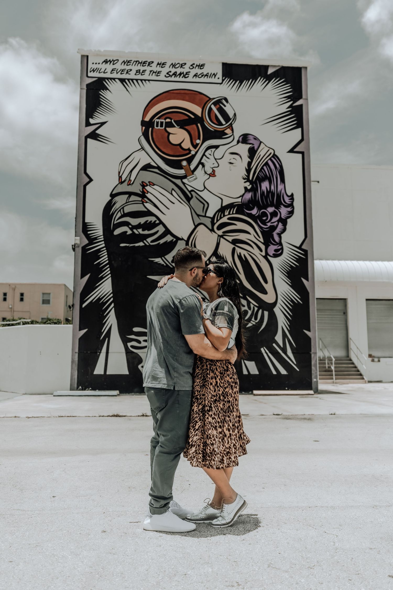 A couple embraces in front of a large street art mural showing an astronaut and figure in a romantic pose.