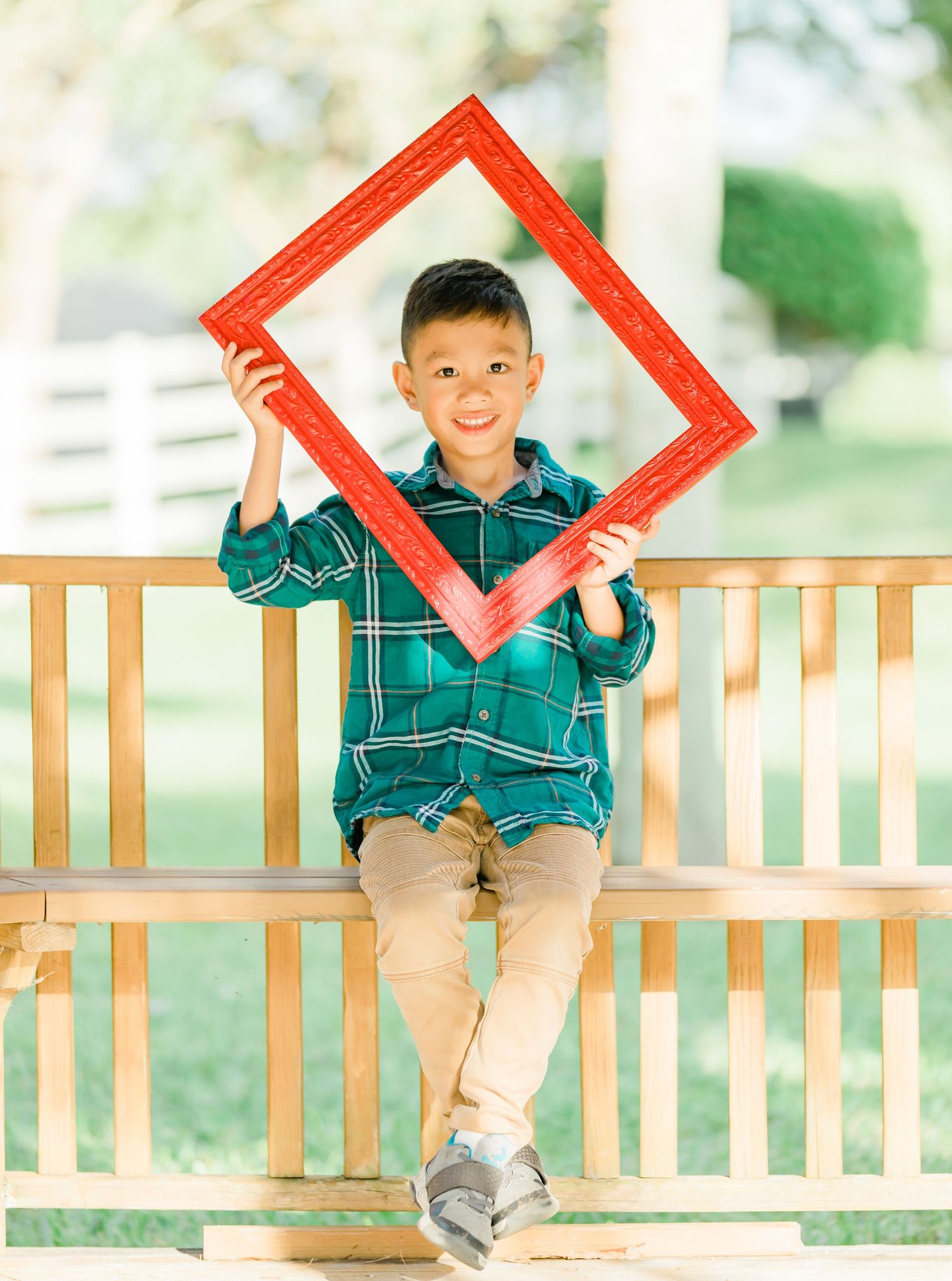 Child in plaid shirt on wooden bench holding large red frame while smiling.
