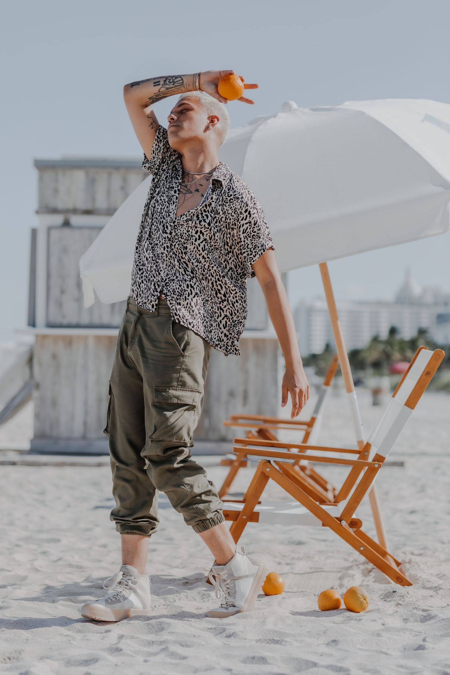 Person in printed shirt and cargo pants posing playfully on beach with oranges.