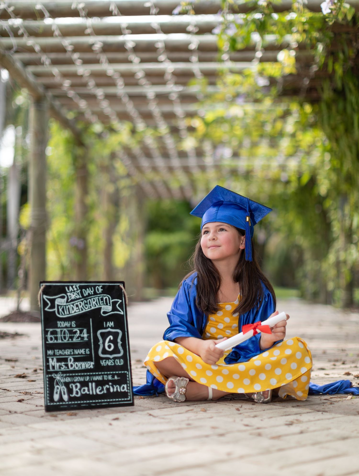 Graduate wearing blue cap and gown sits in garden arbor beside chalkboard sign in polka dot yellow dress.