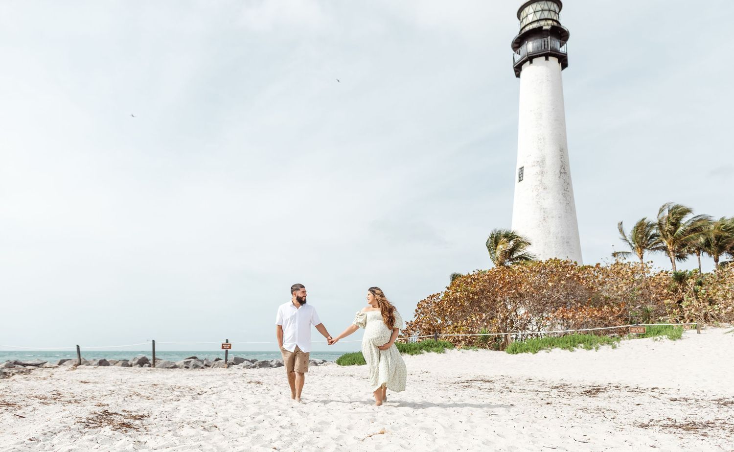 Two people walk hand in hand on a white sandy beach with a tall lighthouse and palm trees in the background.