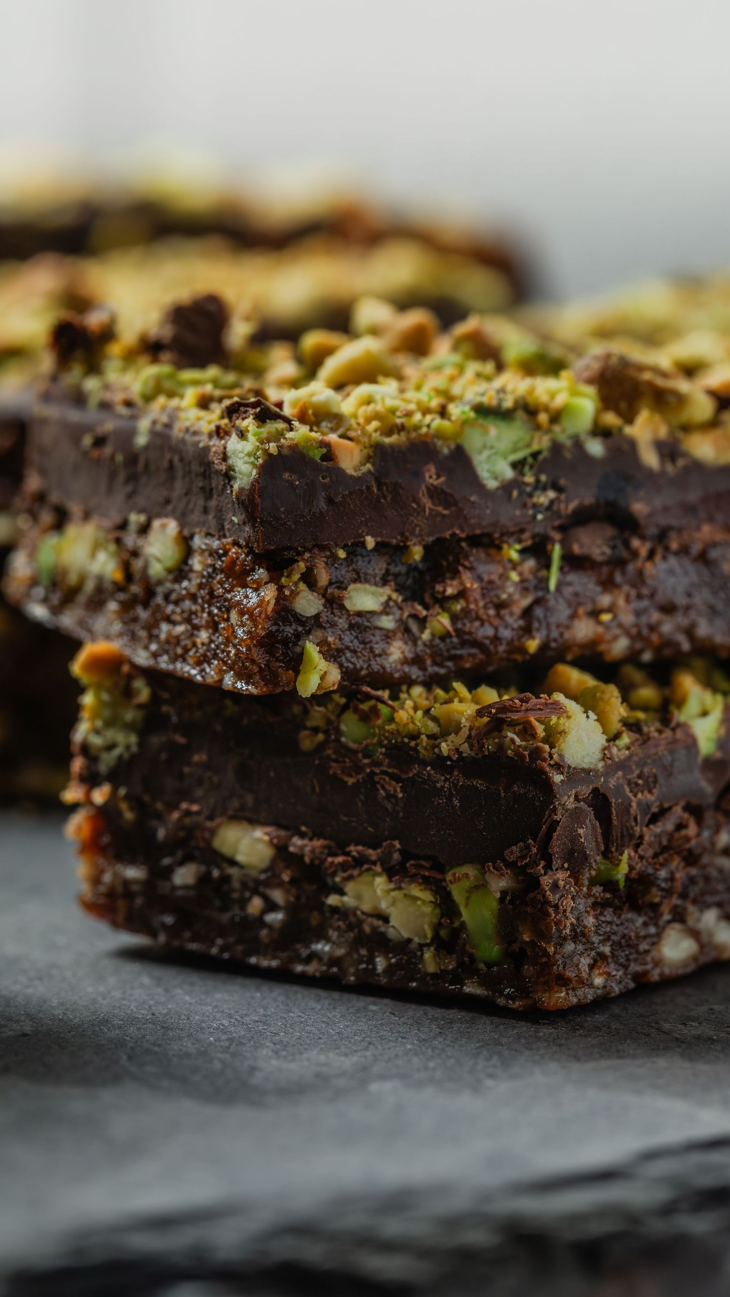 Close-up view of dark chocolate brownies topped with crushed pistachios on a black slate surface.