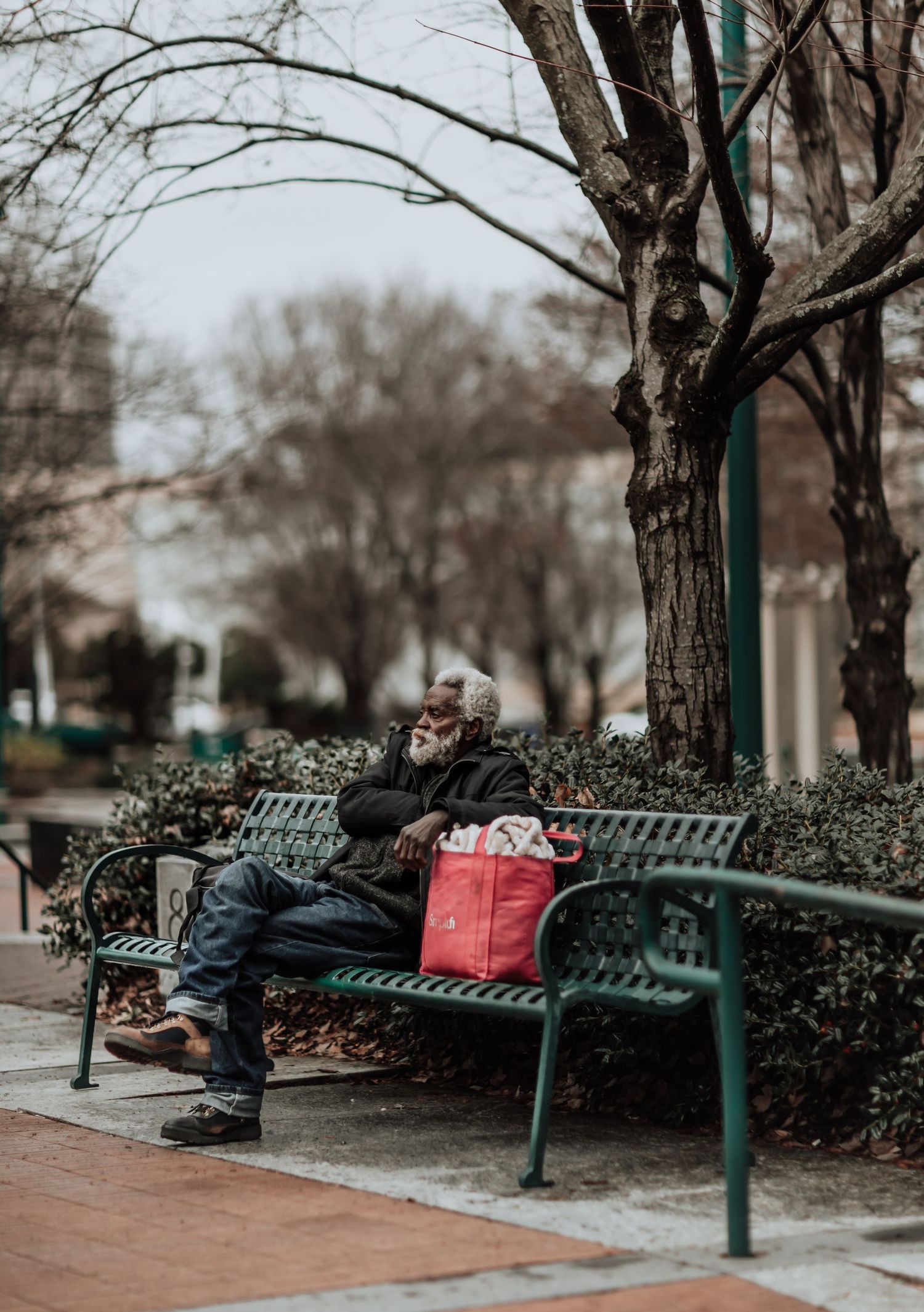 A person rests on a green park bench with shopping bags on a winter day surrounded by bare trees.