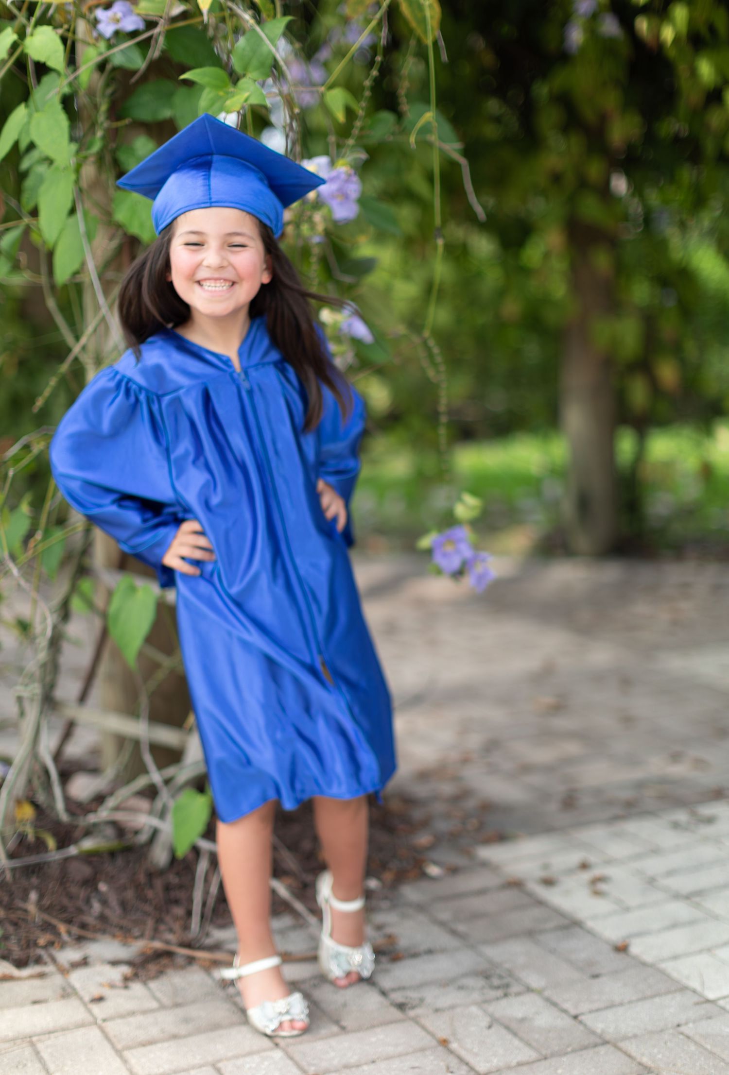 A young graduate in bright blue cap and gown smiles while posing outdoors on a stone path near purple flowers.