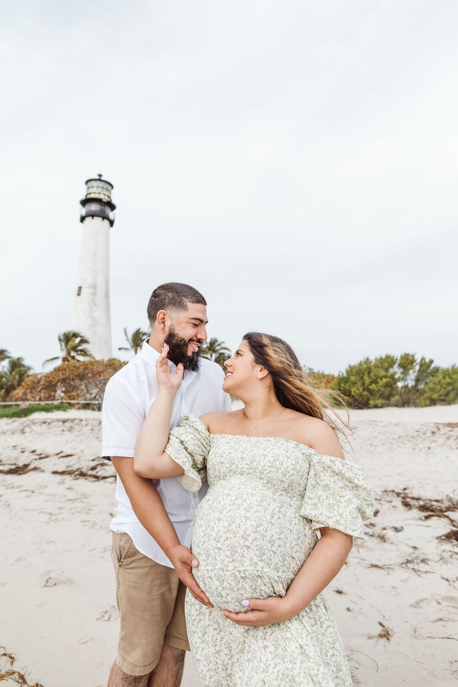 A couple shares an intimate moment during their maternity beach photoshoot near a white lighthouse.