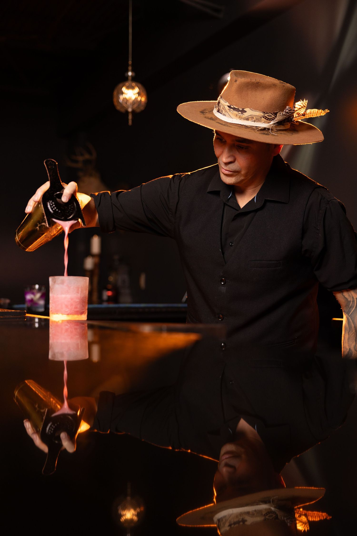 Bartender in black shirt and cowboy hat skillfully pours multiple cocktails simultaneously in dimly lit bar.