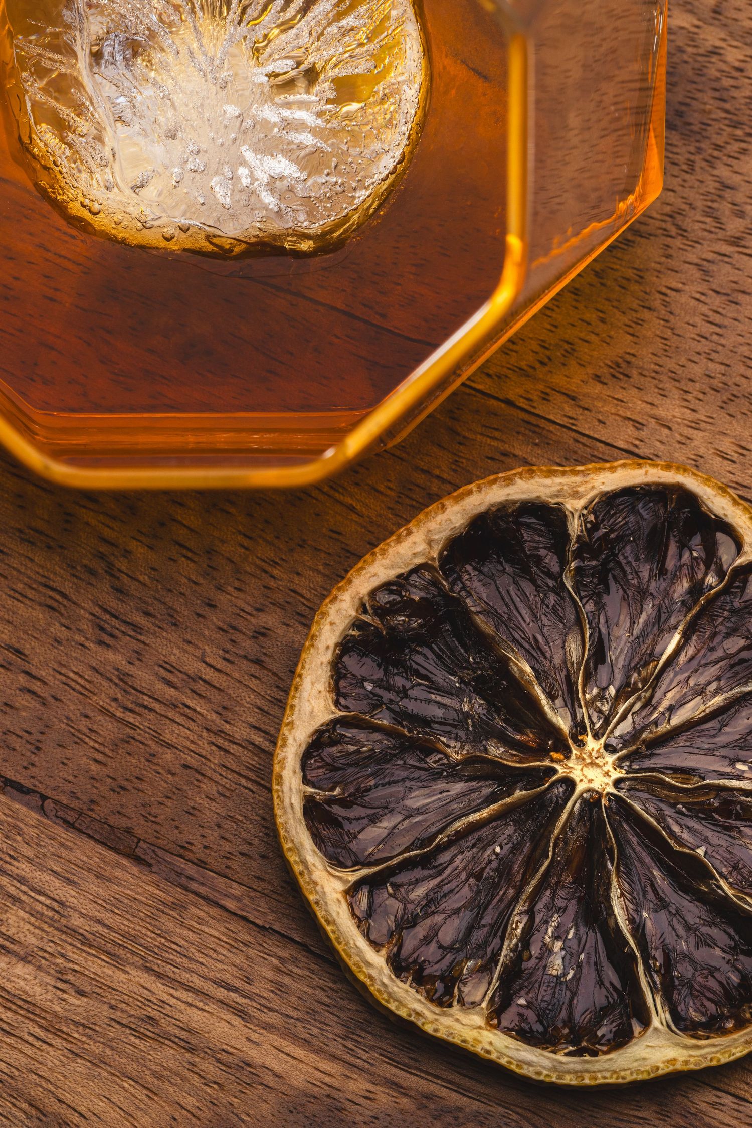 Dried black citrus slice next to a glass of amber liquid on dark wooden surface.