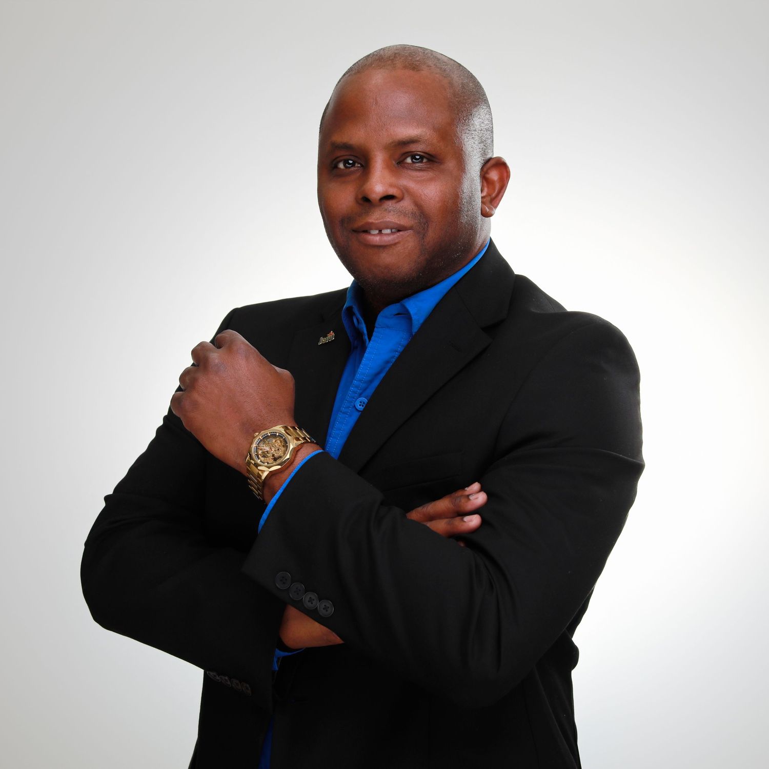 Professional business headshot of person in black suit and blue shirt with crossed arms and gold watch.