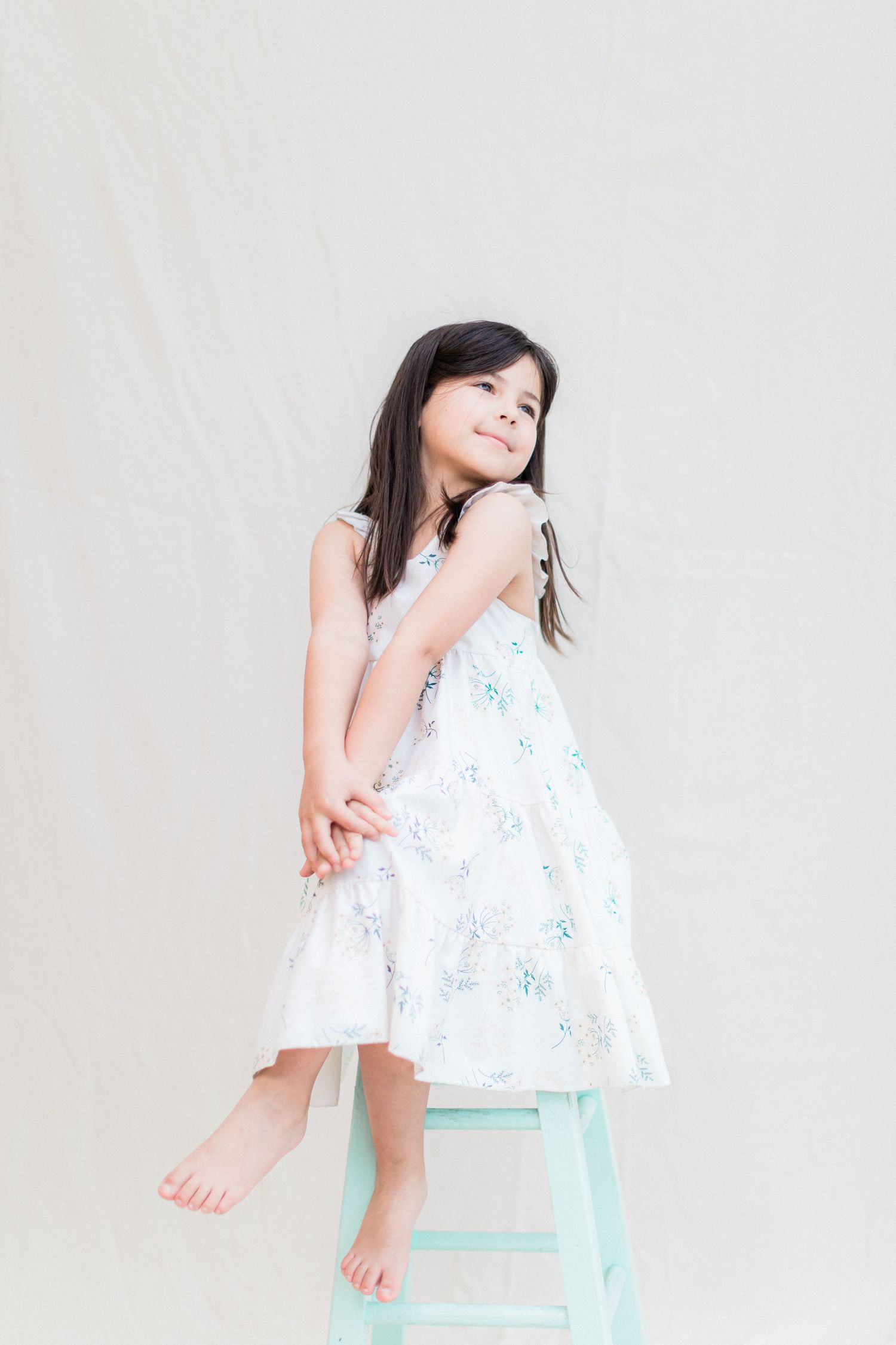 A playful pose on a mint green stool wearing a white floral sundress.