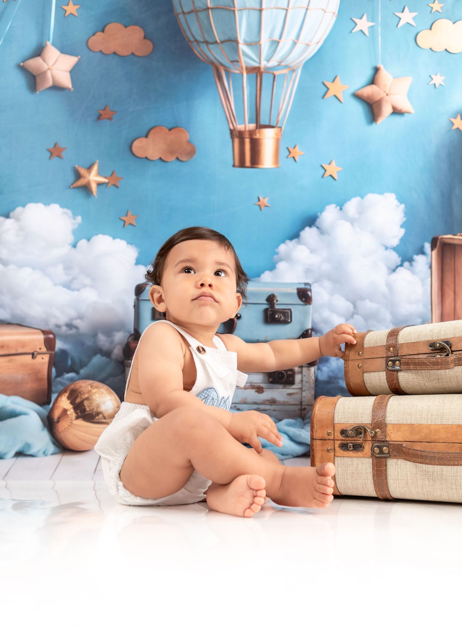 Baby in white outfit sits near vintage luggage with hot air balloon and star decorations overhead.