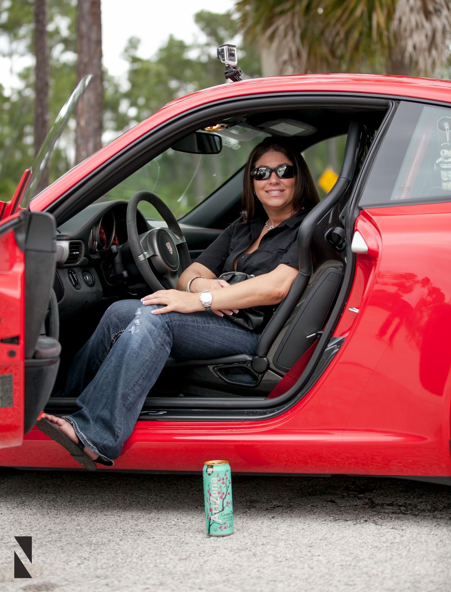 A driver sits in a bright red sports car with a beverage can placed on the ground beside the open door.
