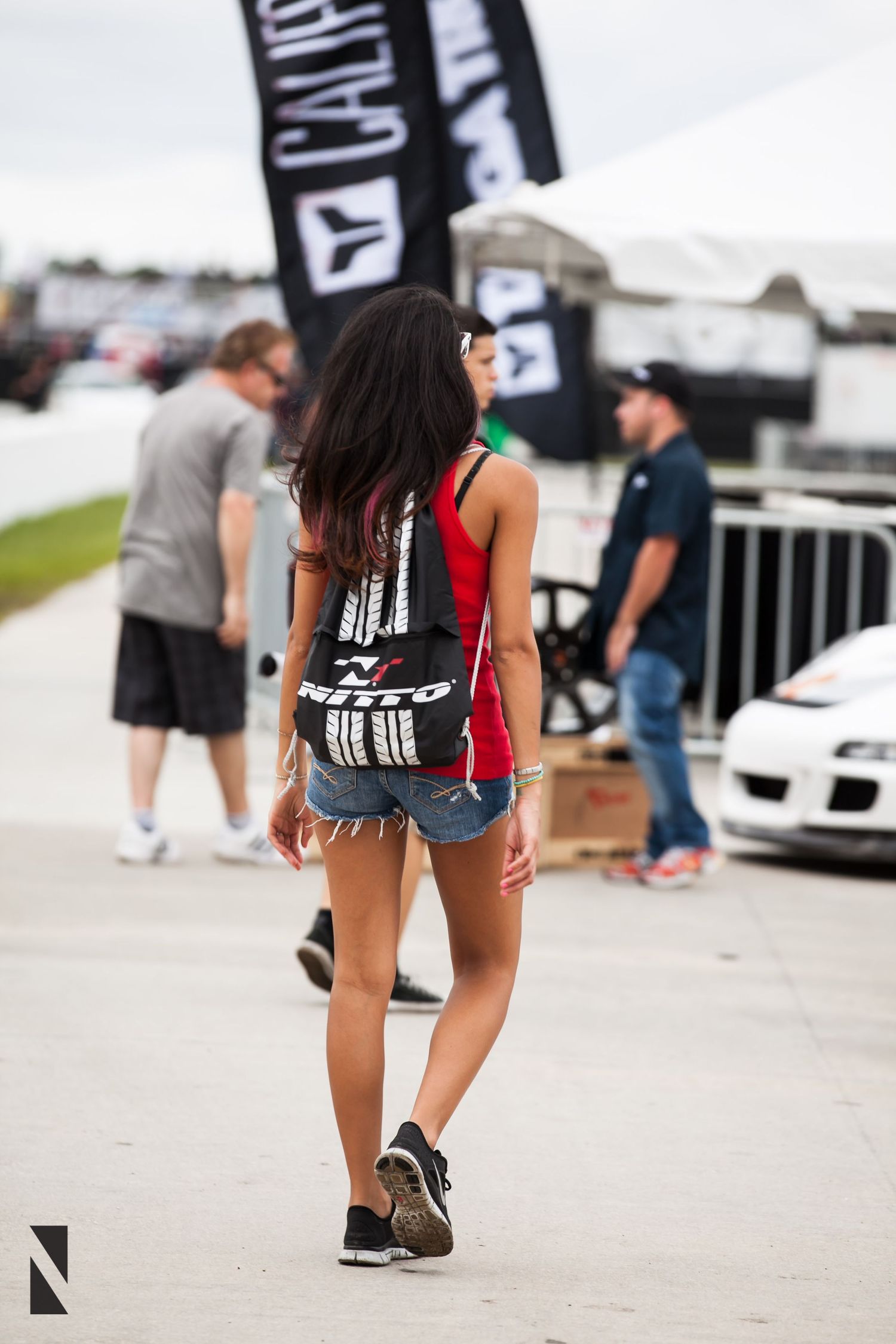 Person in denim shorts and tank top walking through an outdoor car event with black banners and vehicles visible.