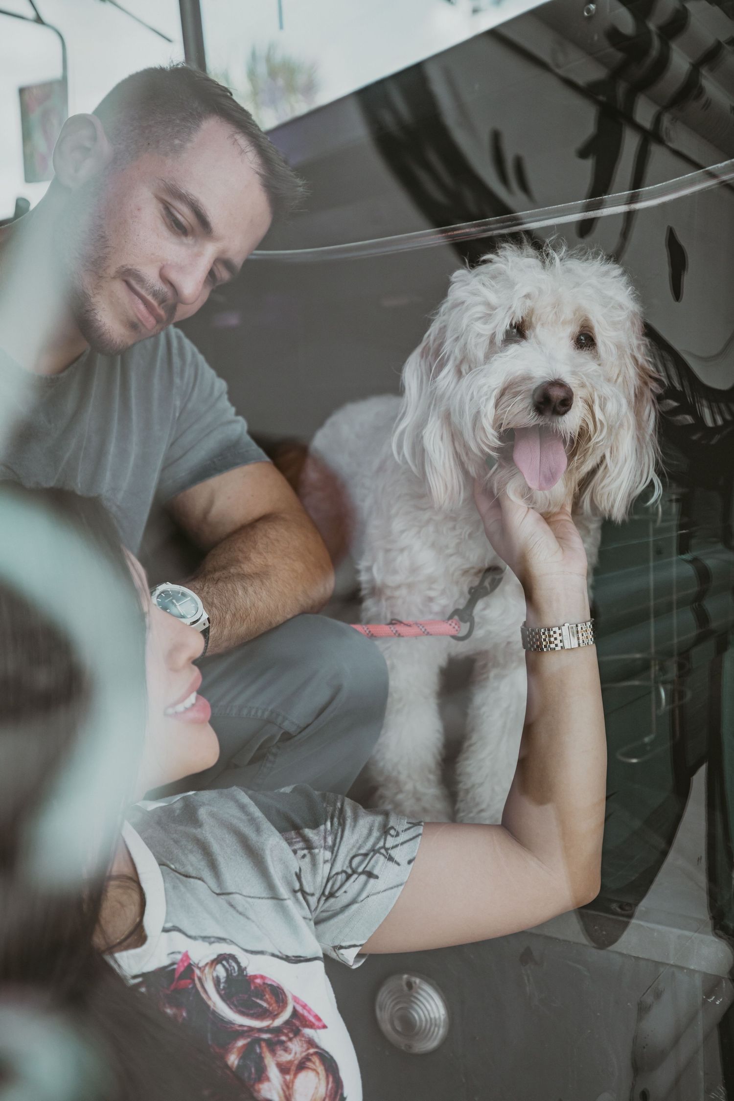 A couple sits with their friendly white dog in what appears to be a vintage vehicle interior.