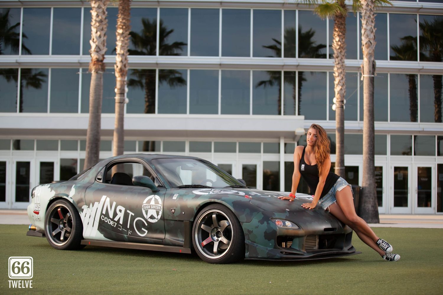 A lowered gray Mazda RX7 sports car with custom decals parked in front of a modern building with palm trees.