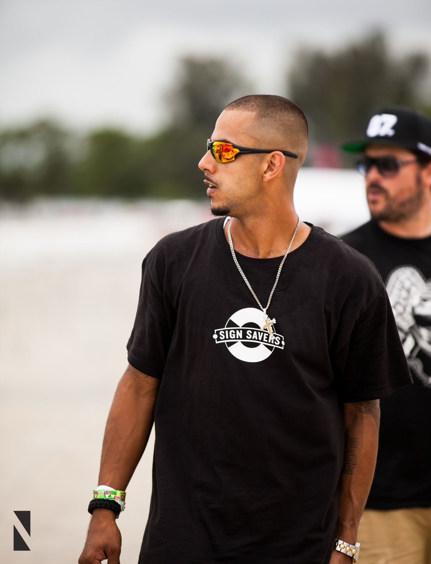 Person wearing black t-shirt and orange sunglasses at outdoor event with someone visible in background.