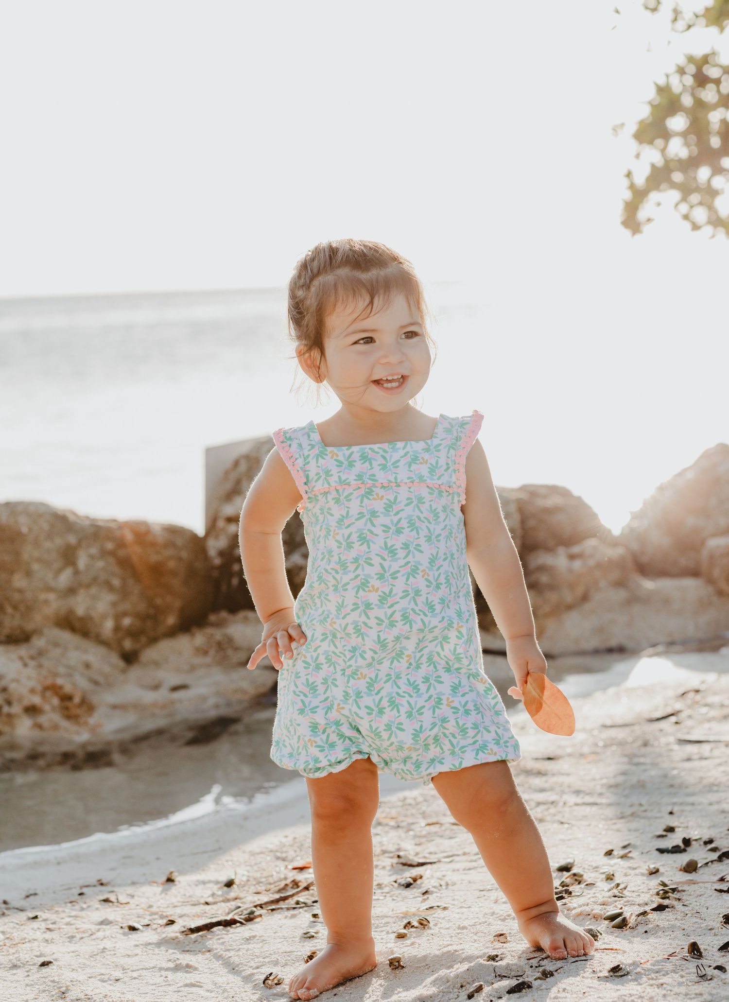 A child in a floral dress plays on a rocky beach during golden hour.