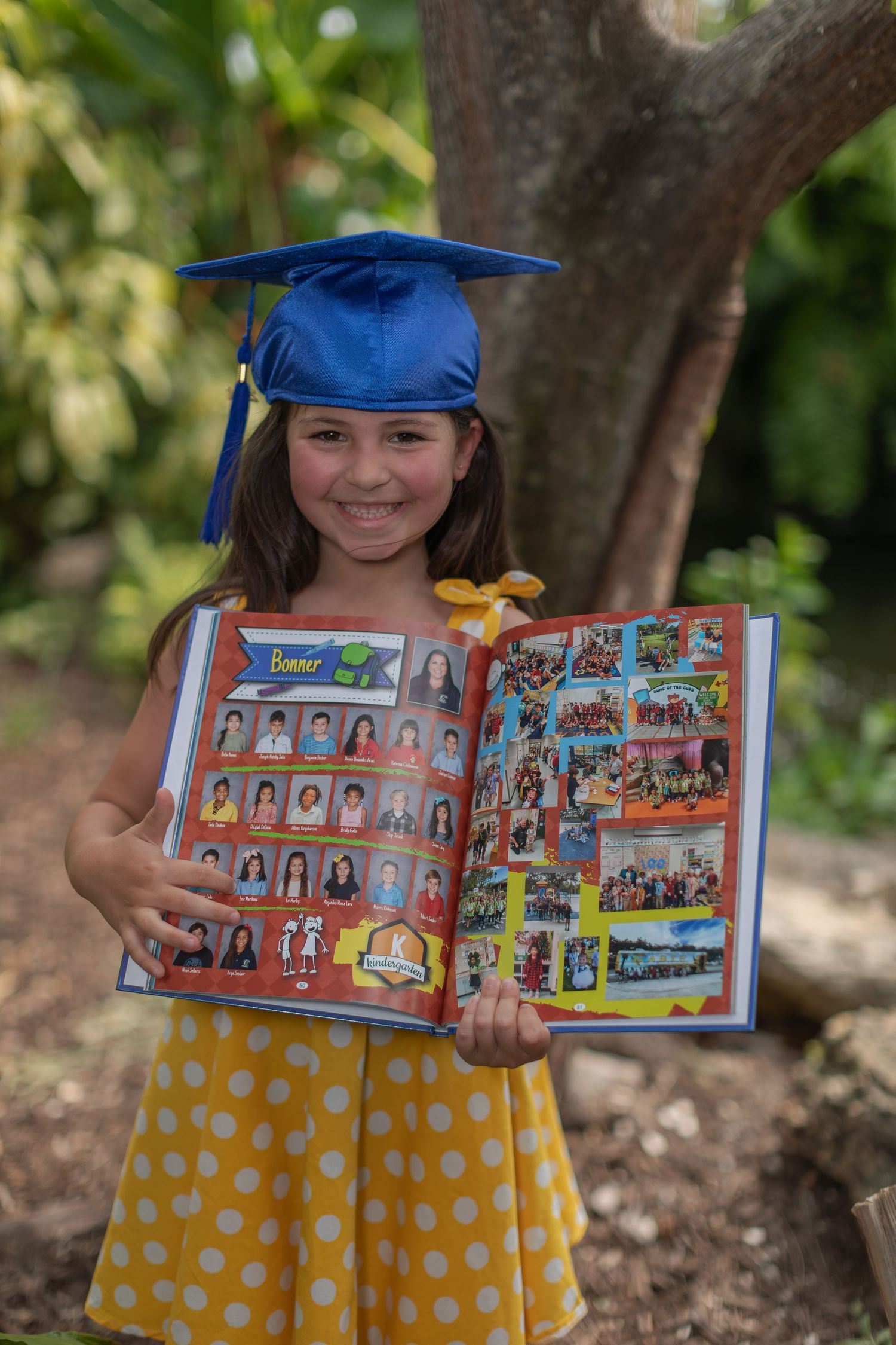 Graduate in blue cap and yellow polka dot dress proudly displays yearbook photos outdoors by a tree.