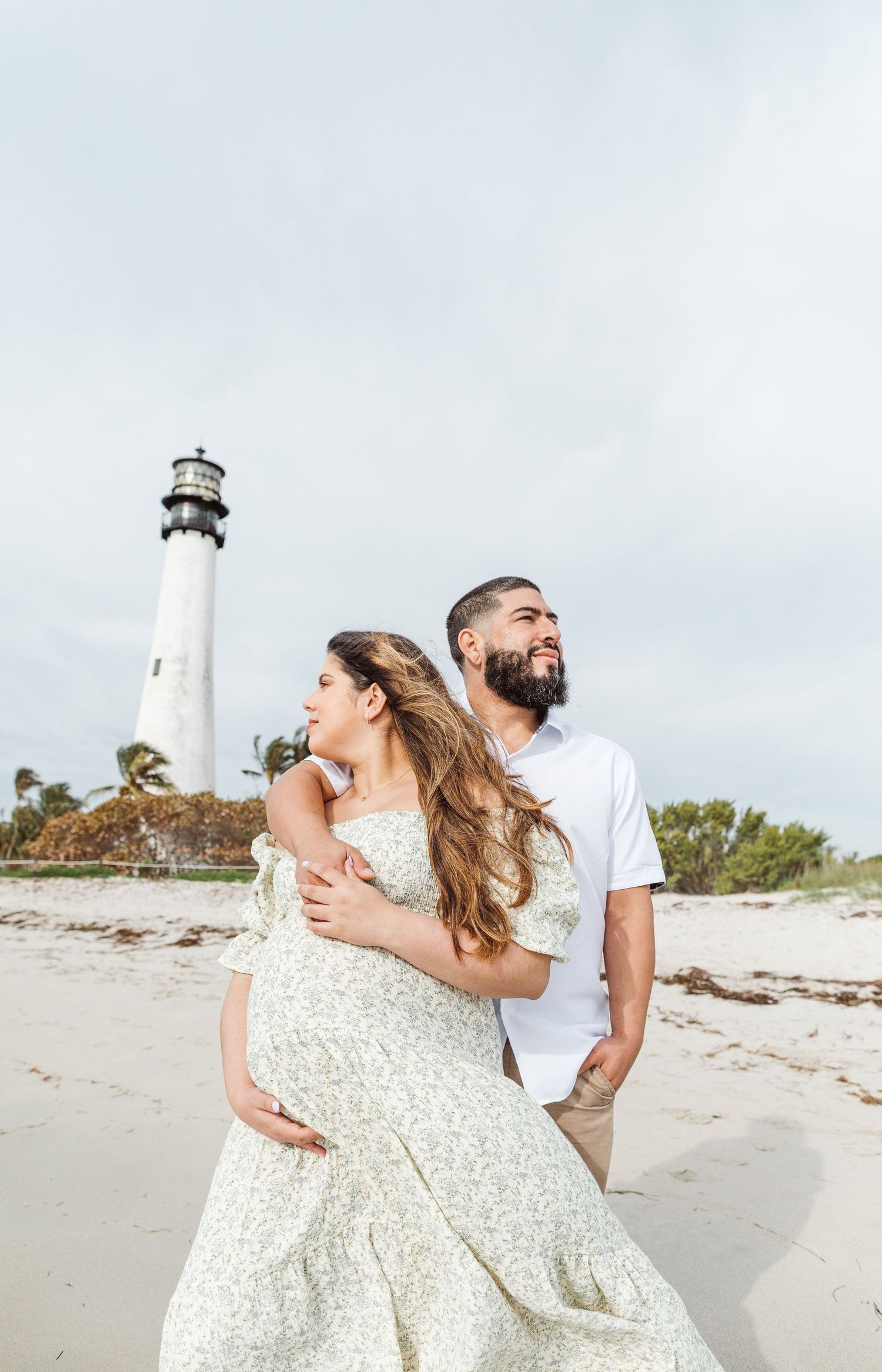 A couple embraces on a sandy beach with a tall white lighthouse in the background on a cloudy day.