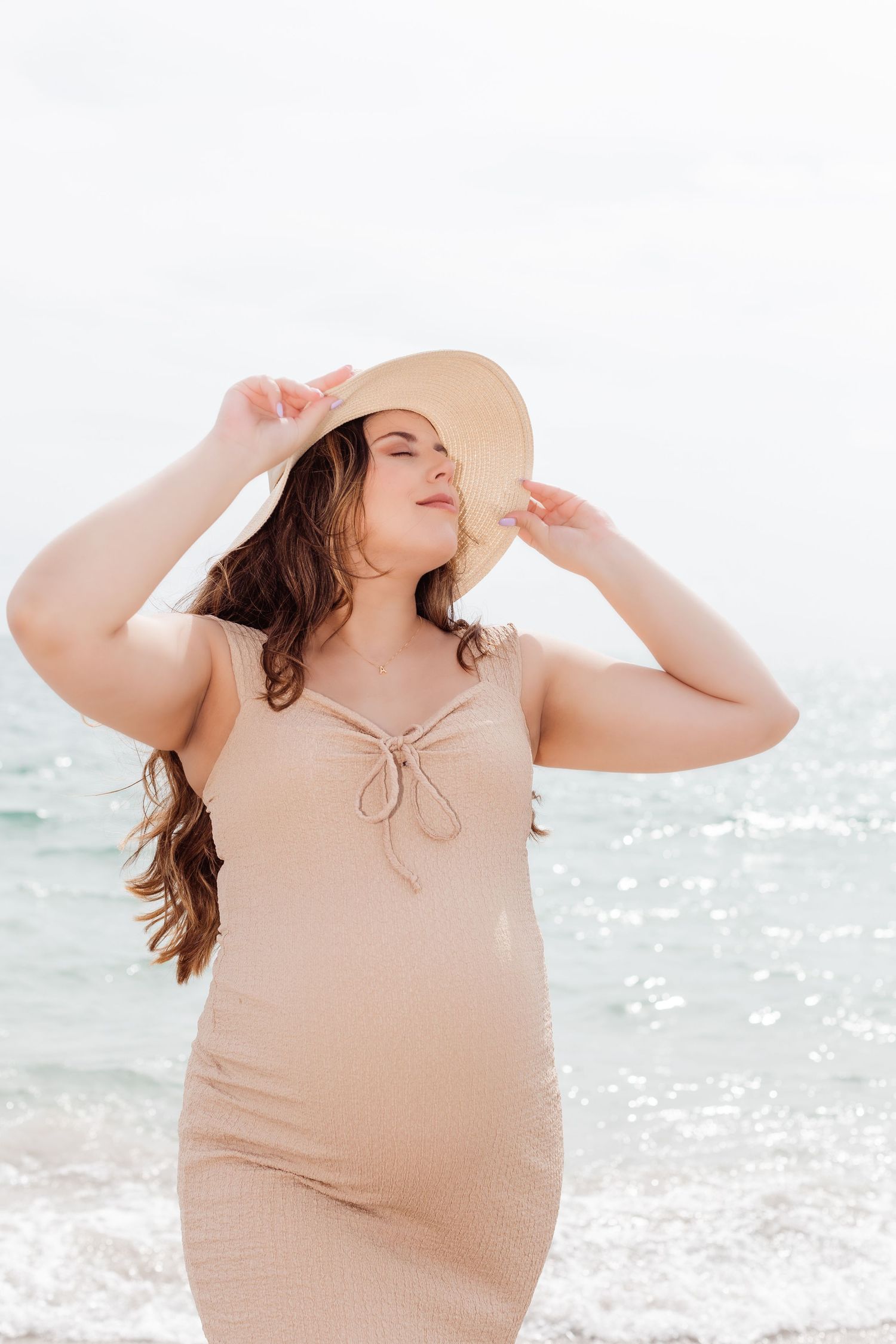 Woman in beige sundress and wide-brimmed hat enjoying a sunny day at the beach with sparkling ocean waves behind her.