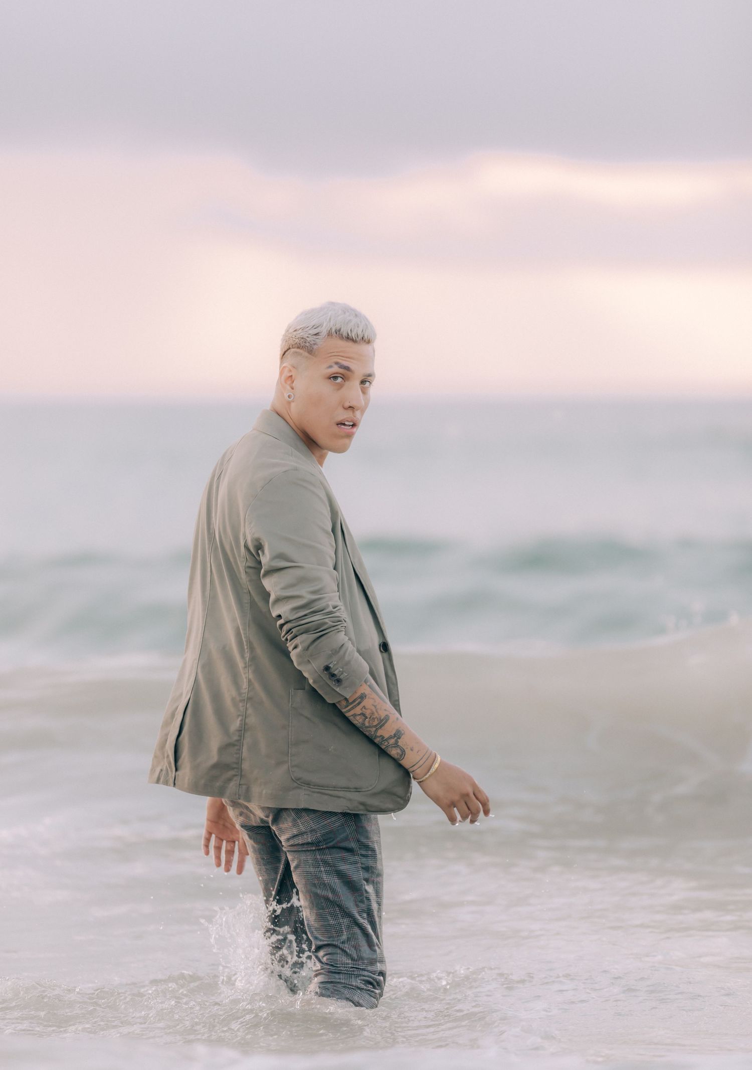 Figure wading in shallow ocean water wearing grey jacket and jeans at dusk.