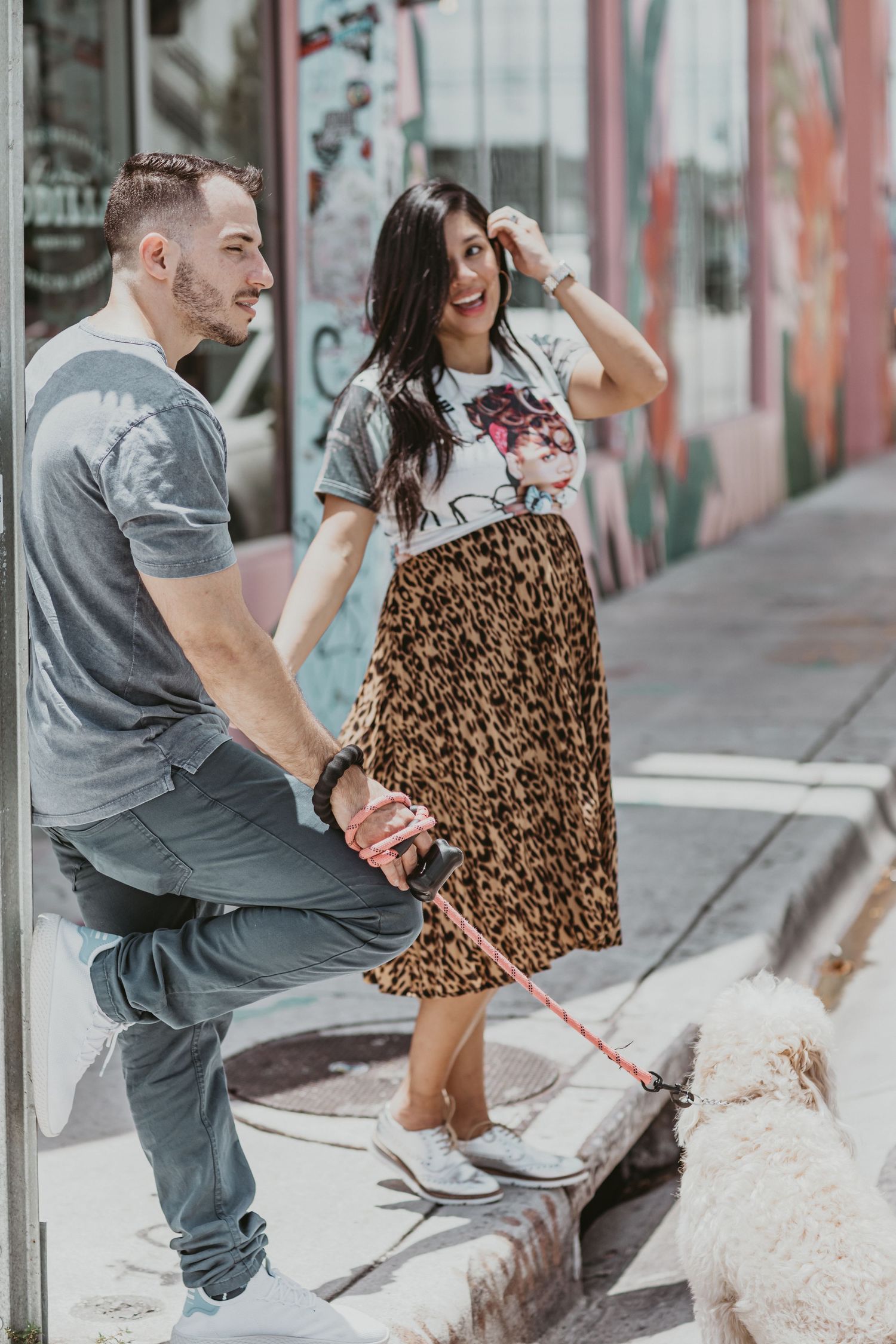 A couple and their white dog pose together against a graffiti wall while wearing casual outfits.