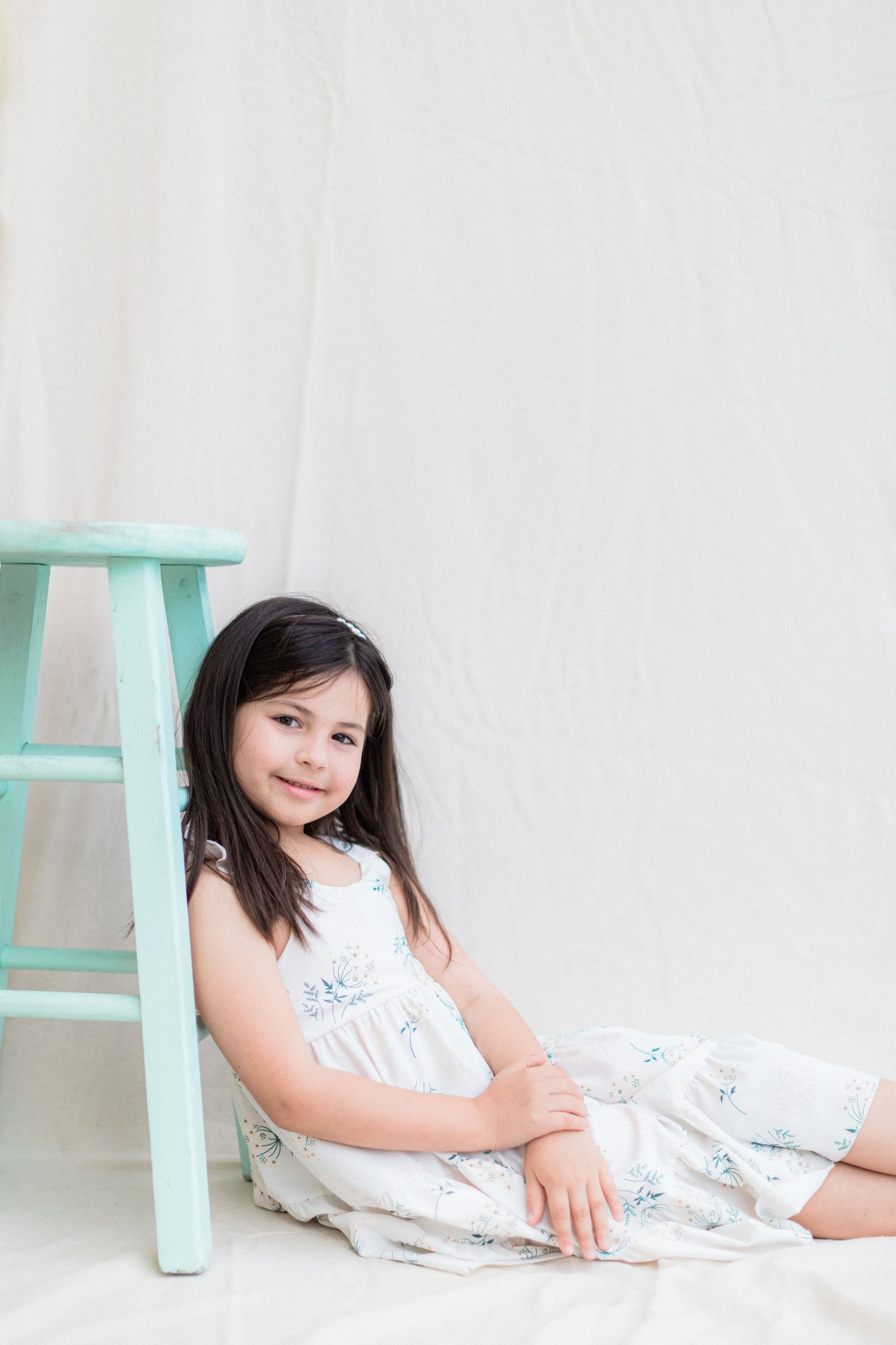A child sits casually on the floor wearing a white floral dress next to a mint green stool.