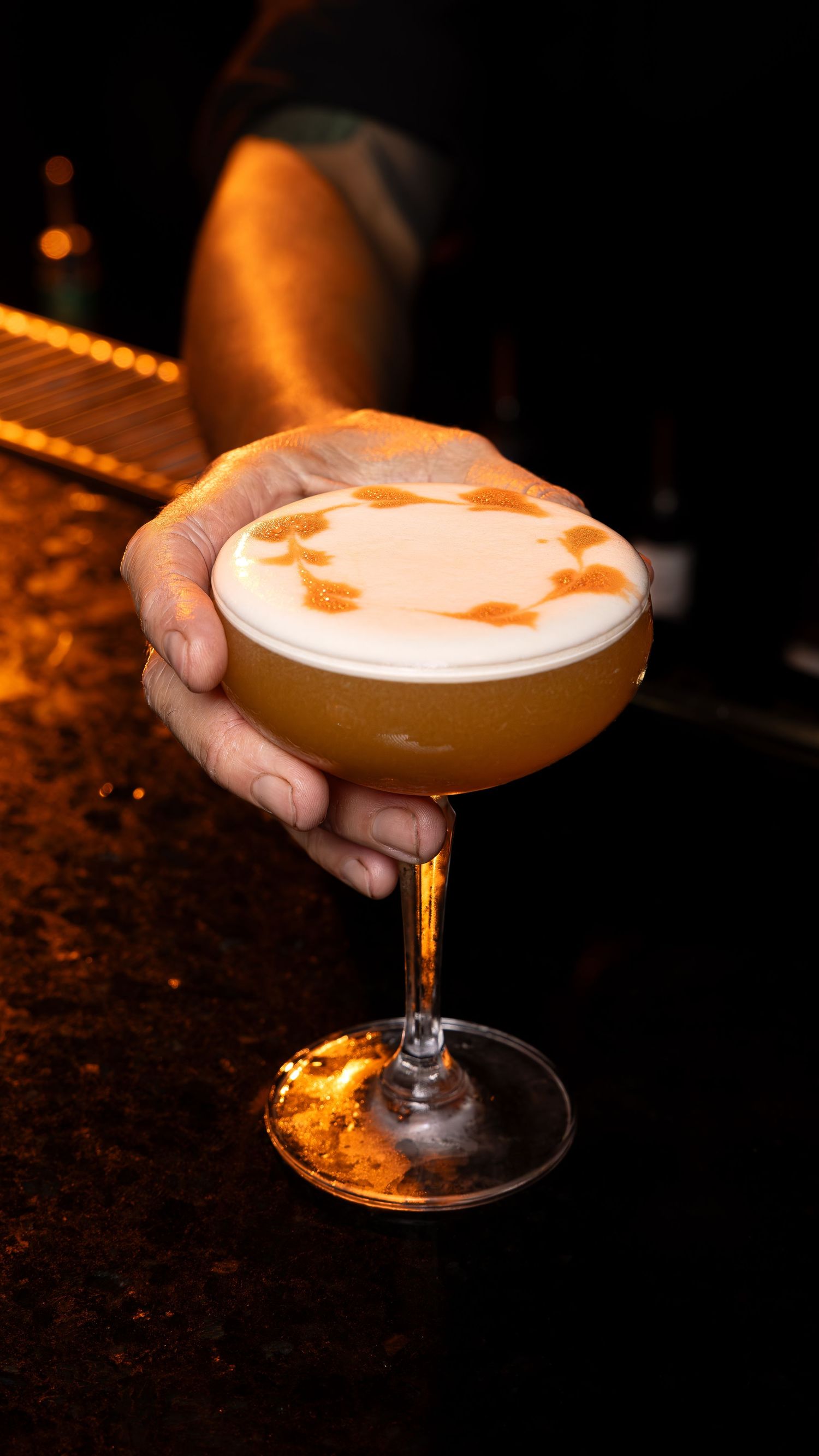 Frothy orange-tinted cocktail served in a coupe glass with artistic foam design on dark wooden surface.