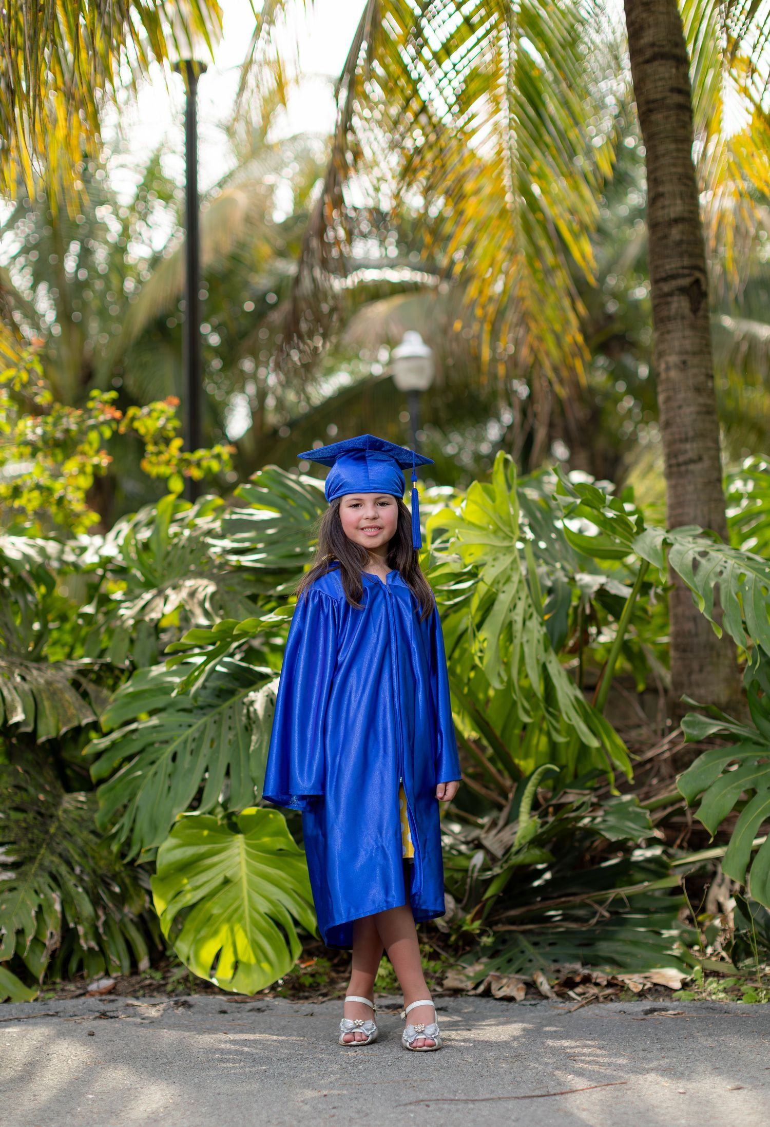 A small figure in bright blue graduation cap and gown stands on a path surrounded by tropical palm fronds and greenery.