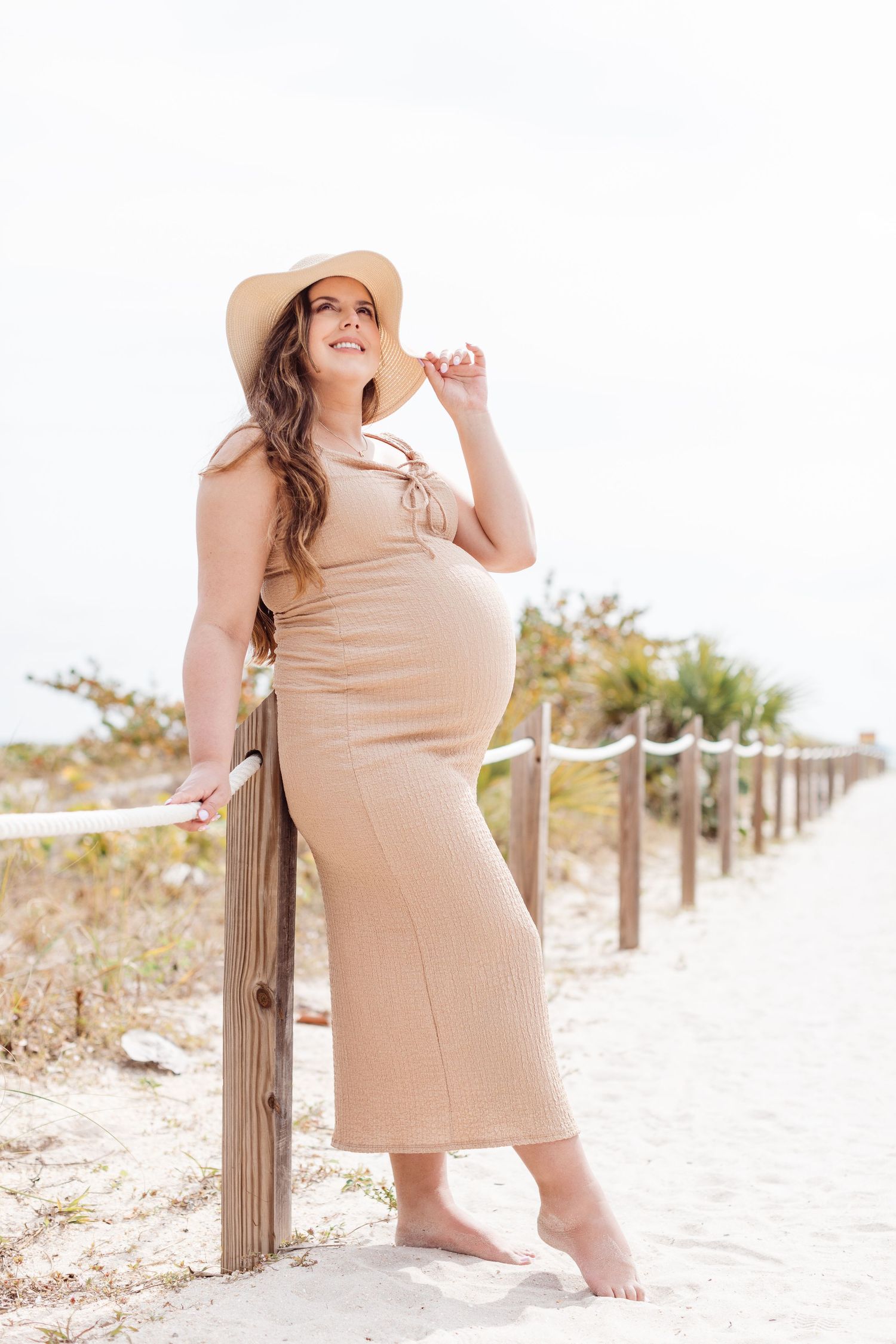 Expectant mother in beige dress and hat poses barefoot against wooden railing on sandy beach path during sunset.