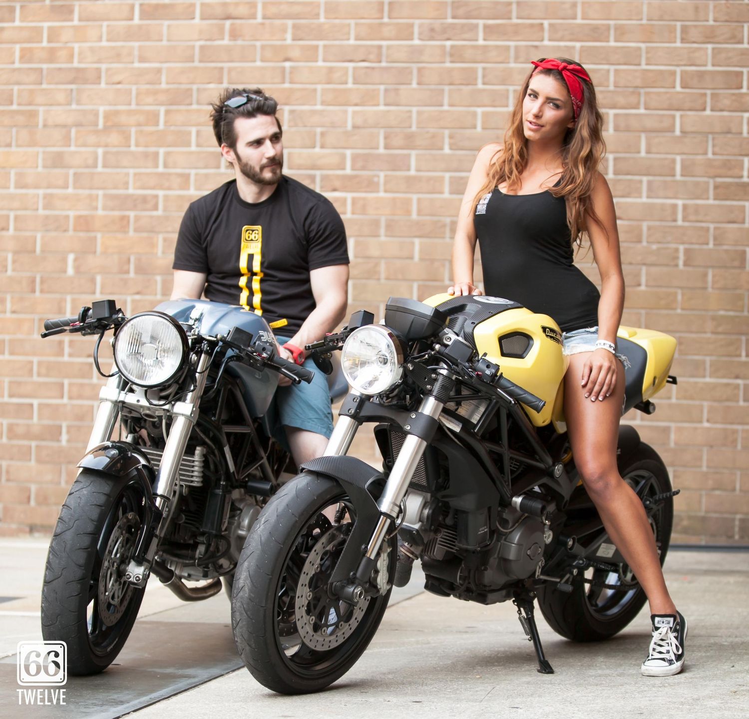 Two motorcyclists pose with vintage cafe racer style motorcycles against a brick wall background.