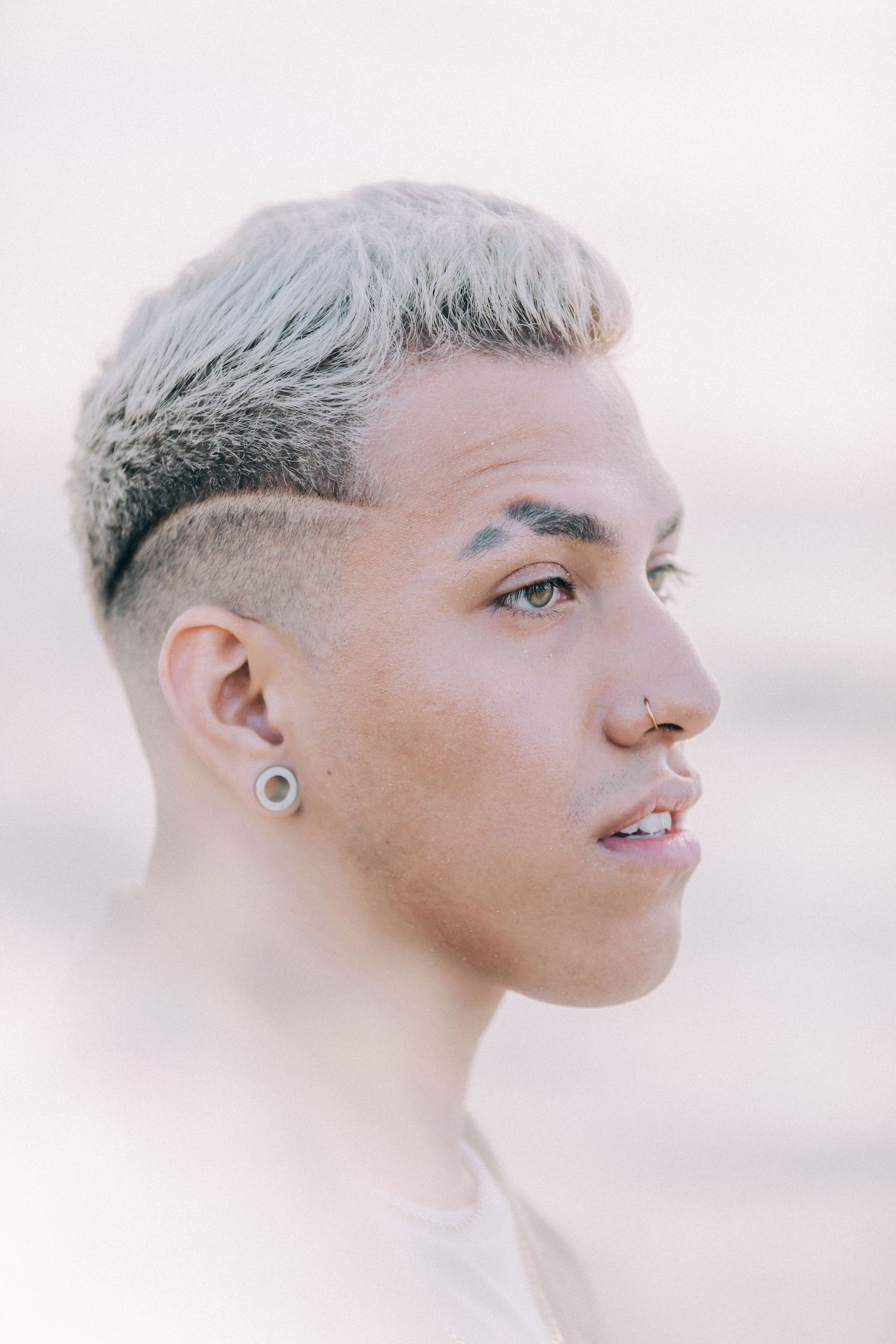 Profile portrait of person with platinum blonde hair and ear piercing against soft pink background.