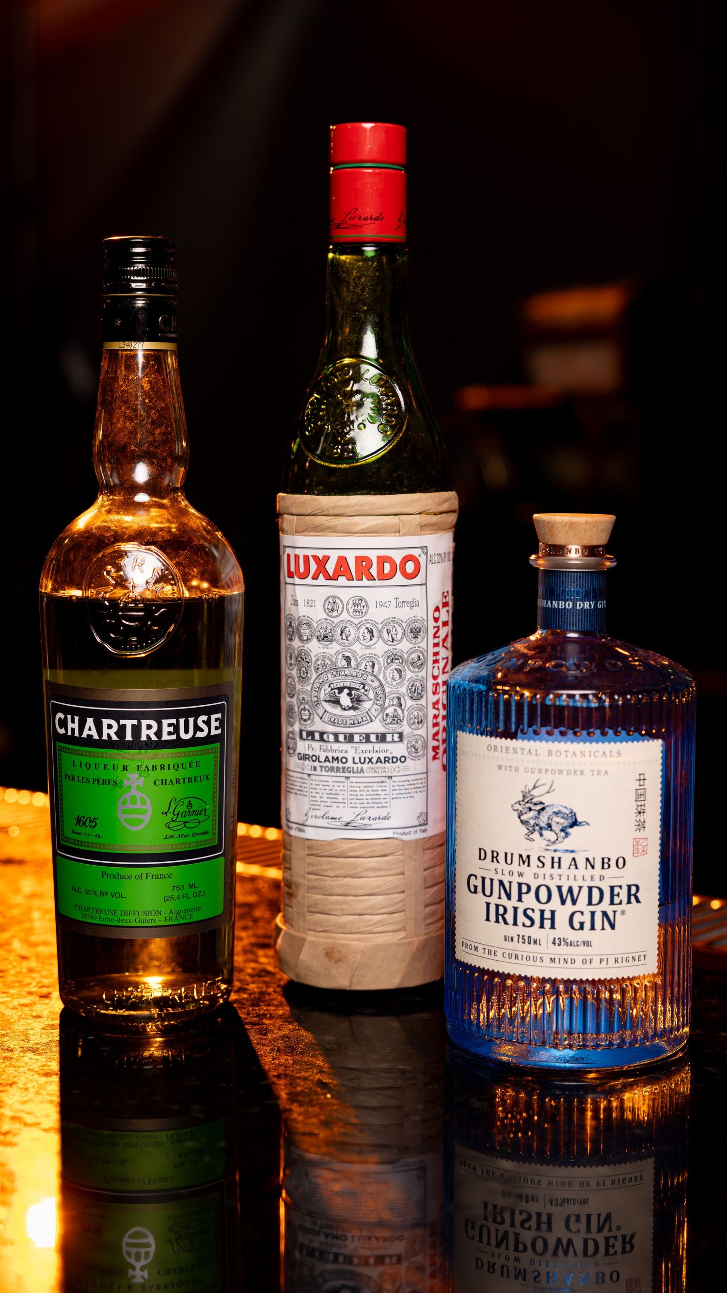 Three craft liquor bottles including green Chartreuse, Luxardo liqueur, and Drumshanbo Gunpowder Irish Gin on reflective surface.