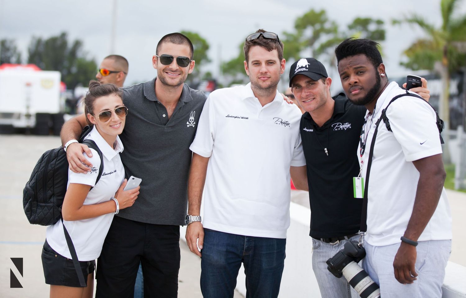 A group of five people wearing casual attire pose together outdoors with palm trees visible in the background.
