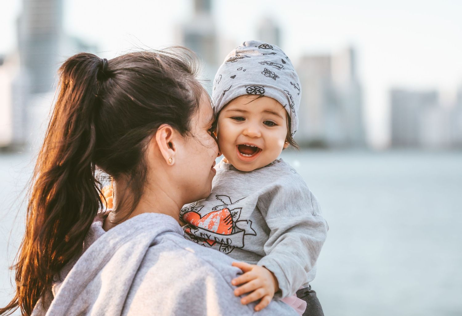 A joyful baby wearing a gray winter hat laughs while being held against a city waterfront backdrop.