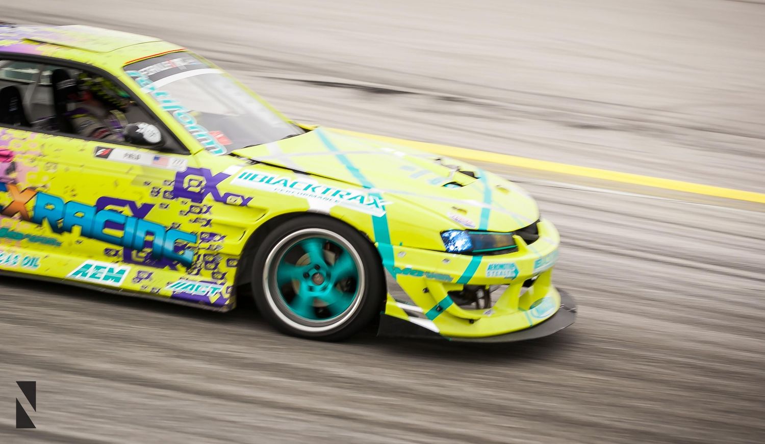 Yellow and turquoise drift car with custom body kit sliding sideways on track during competition.