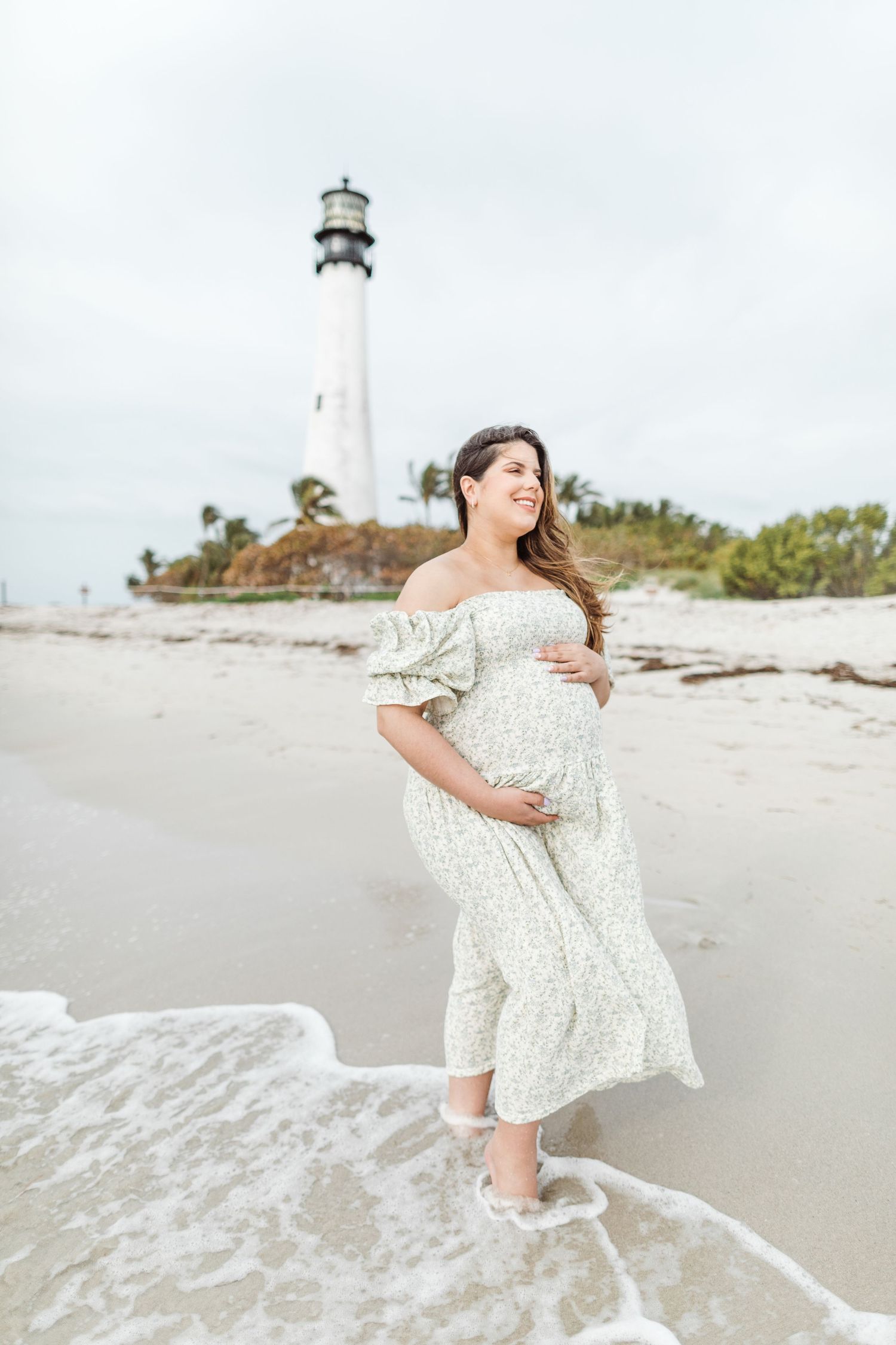 A maternity photo shoot with waves lapping at feet on a beach with a lighthouse in the background.