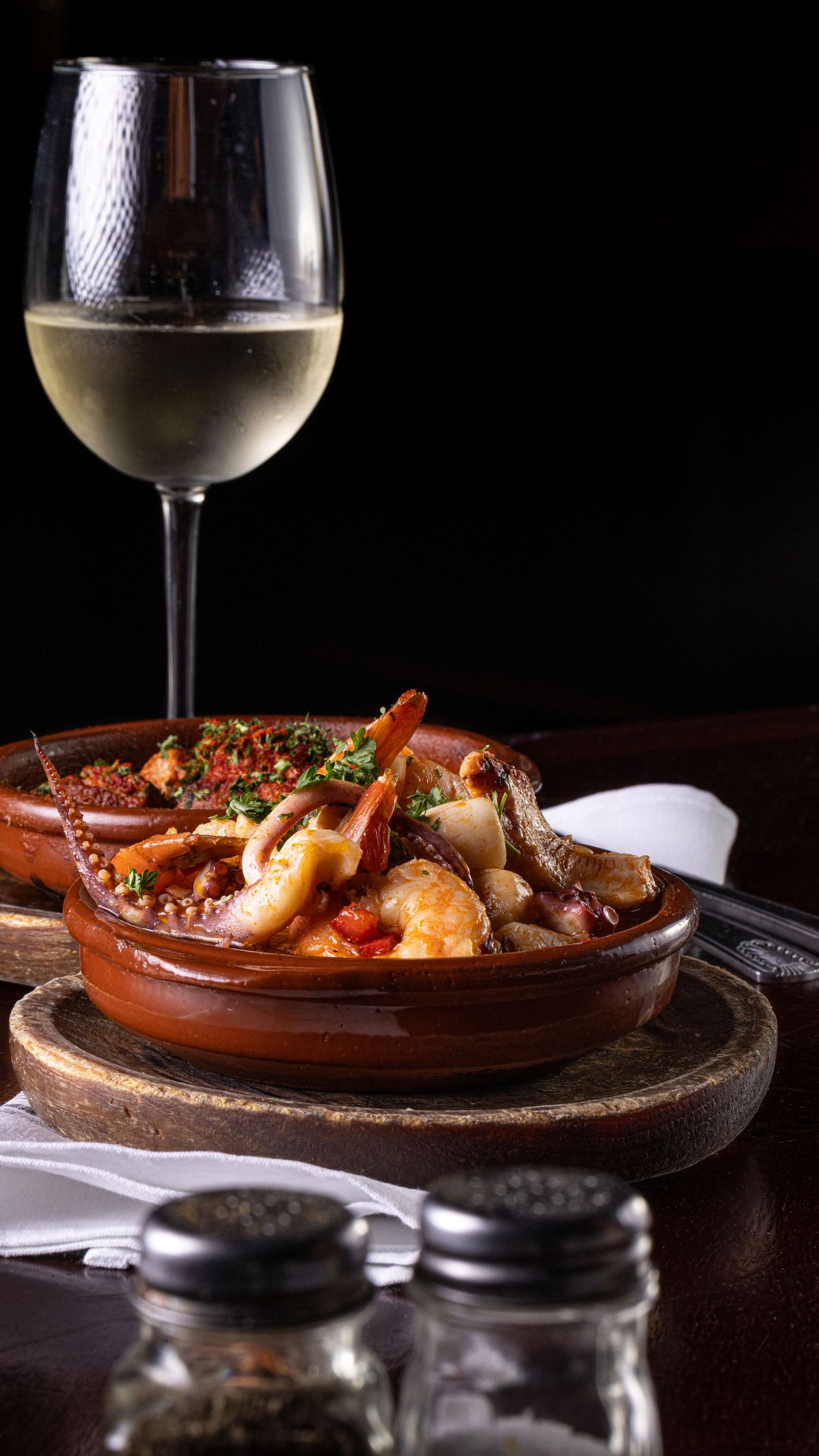 A glass of white wine alongside a terracotta dish of seafood stew on a wooden board with salt and pepper shakers.