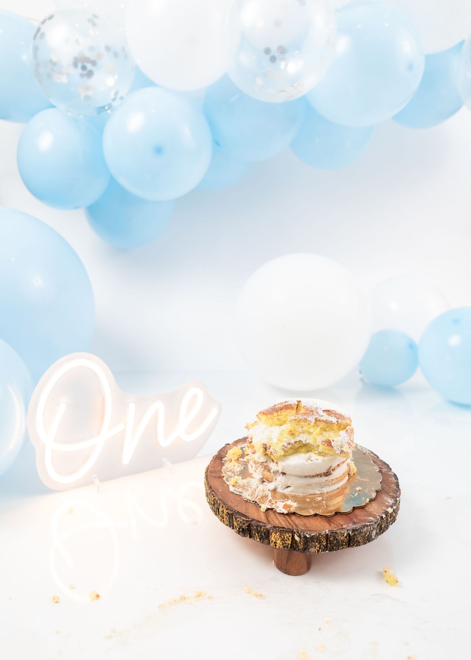 Rustic cake display with 'One' neon sign and blue balloon arch backdrop.