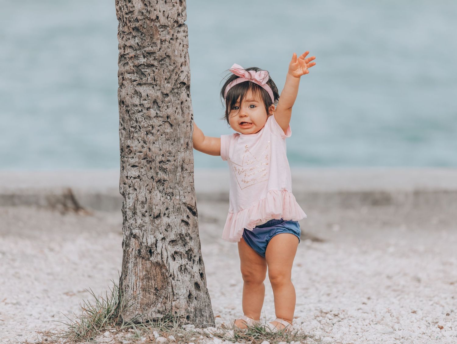 Small child wearing a pink top and denim shorts stands by a tree trunk while raising one arm on a beach.