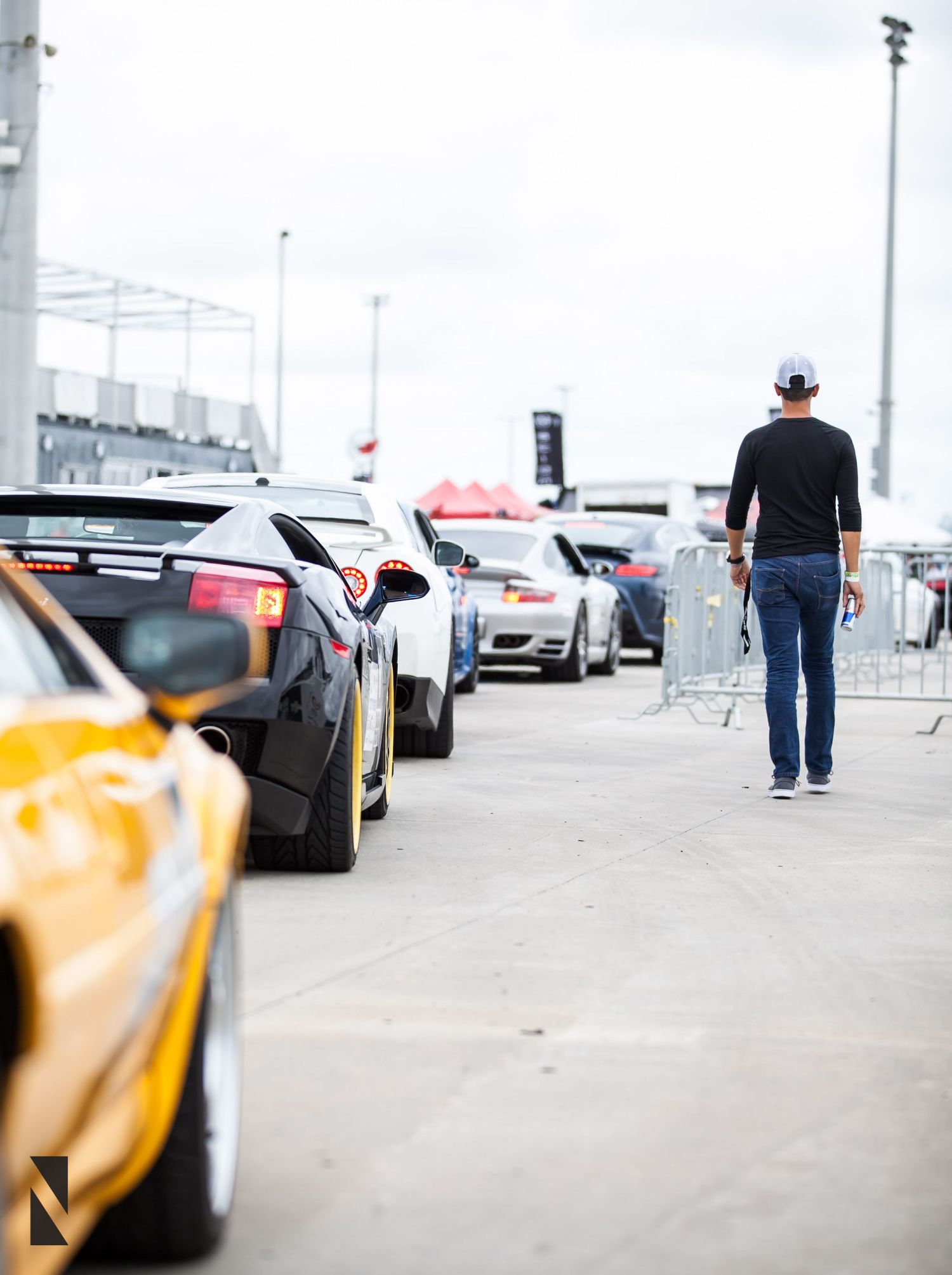 Someone in jeans and cowboy hat walks past line of luxury sports cars and exotic vehicles parked at racing event.